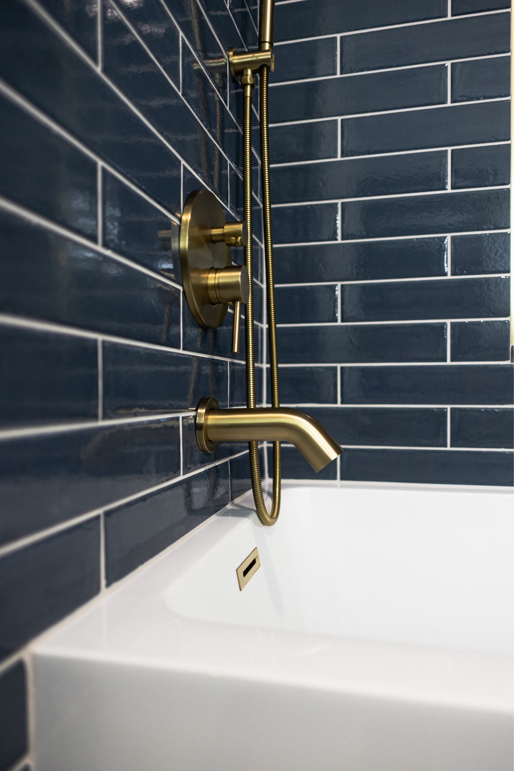 Brass shower fixture with white tub and dark blue subway tile walls