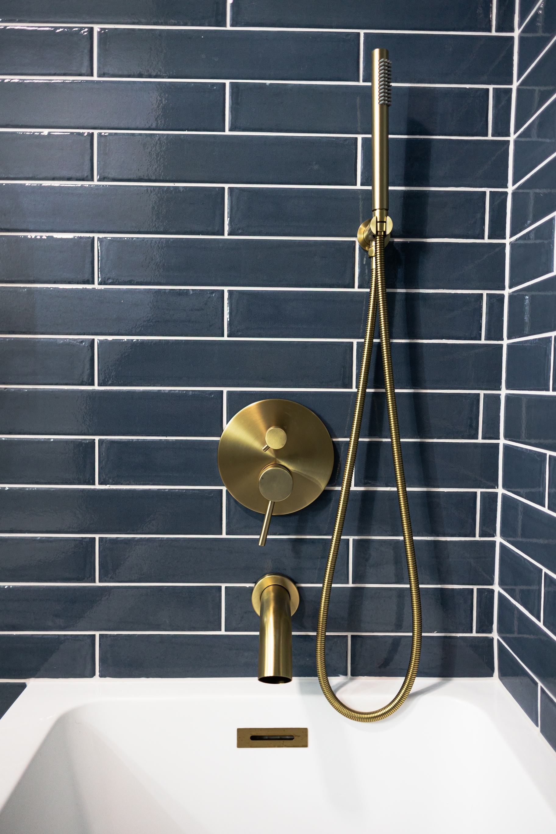 Brass shower fixture with handheld wand over a white tub against dark blue tile walls