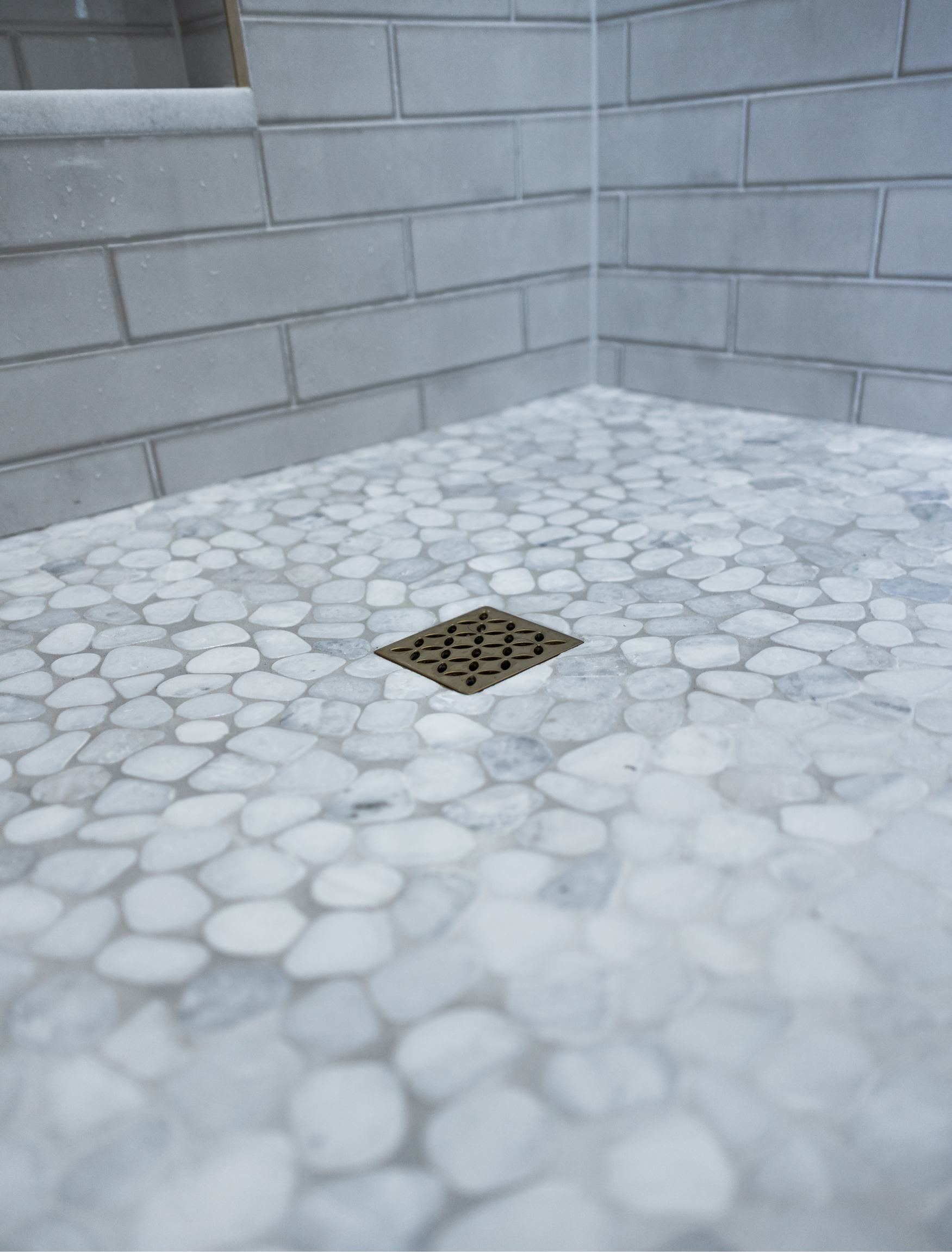 Small square floor drain in a light gray tiled shower corner with pebble-pattern flooring