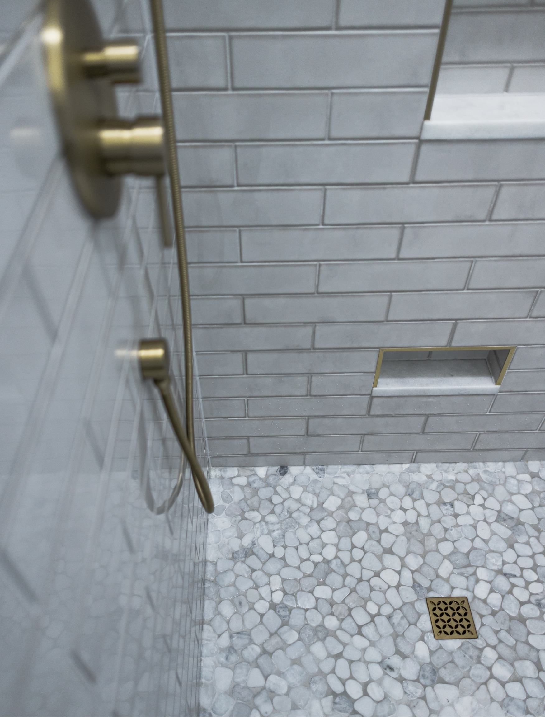 Tiled shower corner with brass fixtures, gray brick walls, pebble floor, and a drain.