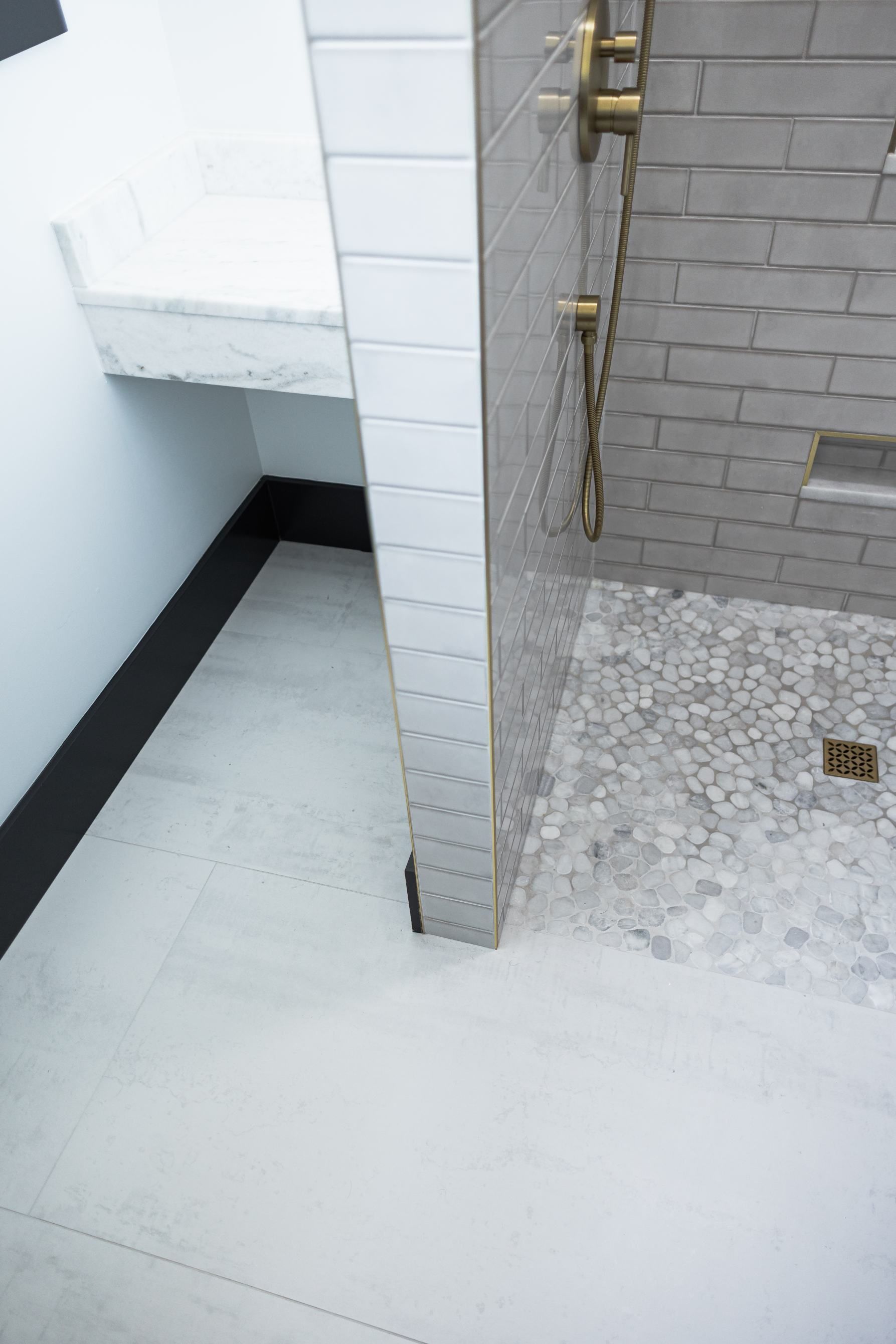 Small shower area with white and gray tiles, brass fixtures, and pebble flooring beside a narrow stairwell.