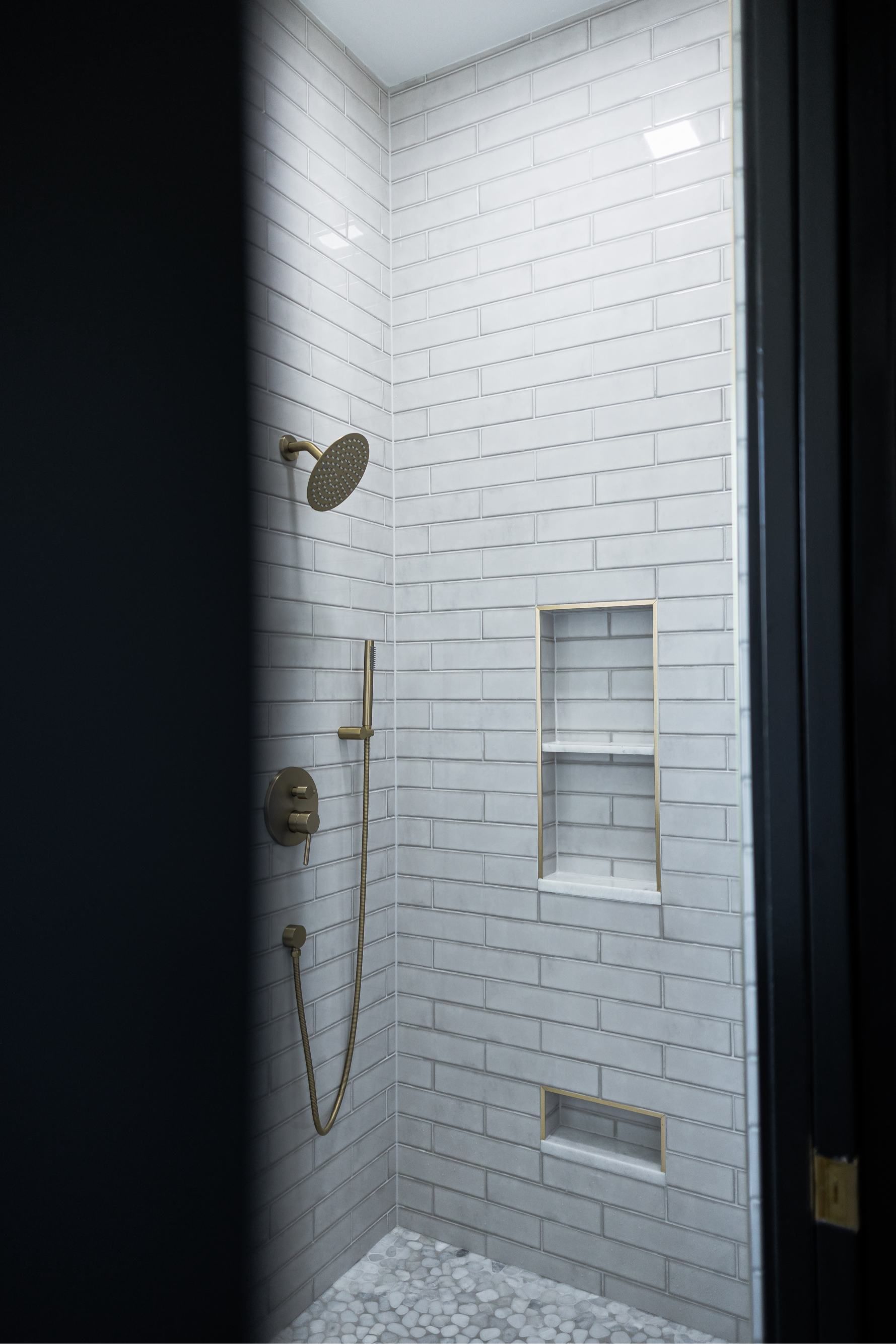 White-tiled shower stall with built-in shelves, handheld showerhead, and black door frame