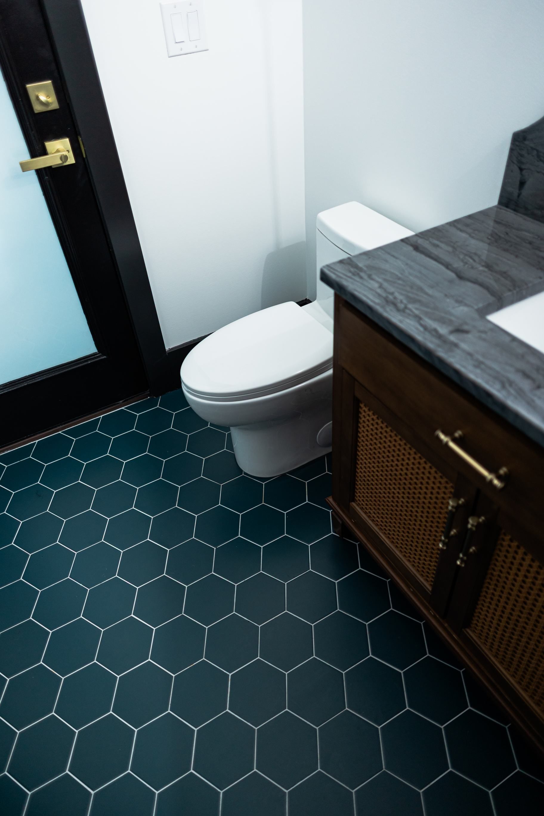 Small bathroom with white toilet, dark hexagon floor tile, and wood vanity with gray countertop.