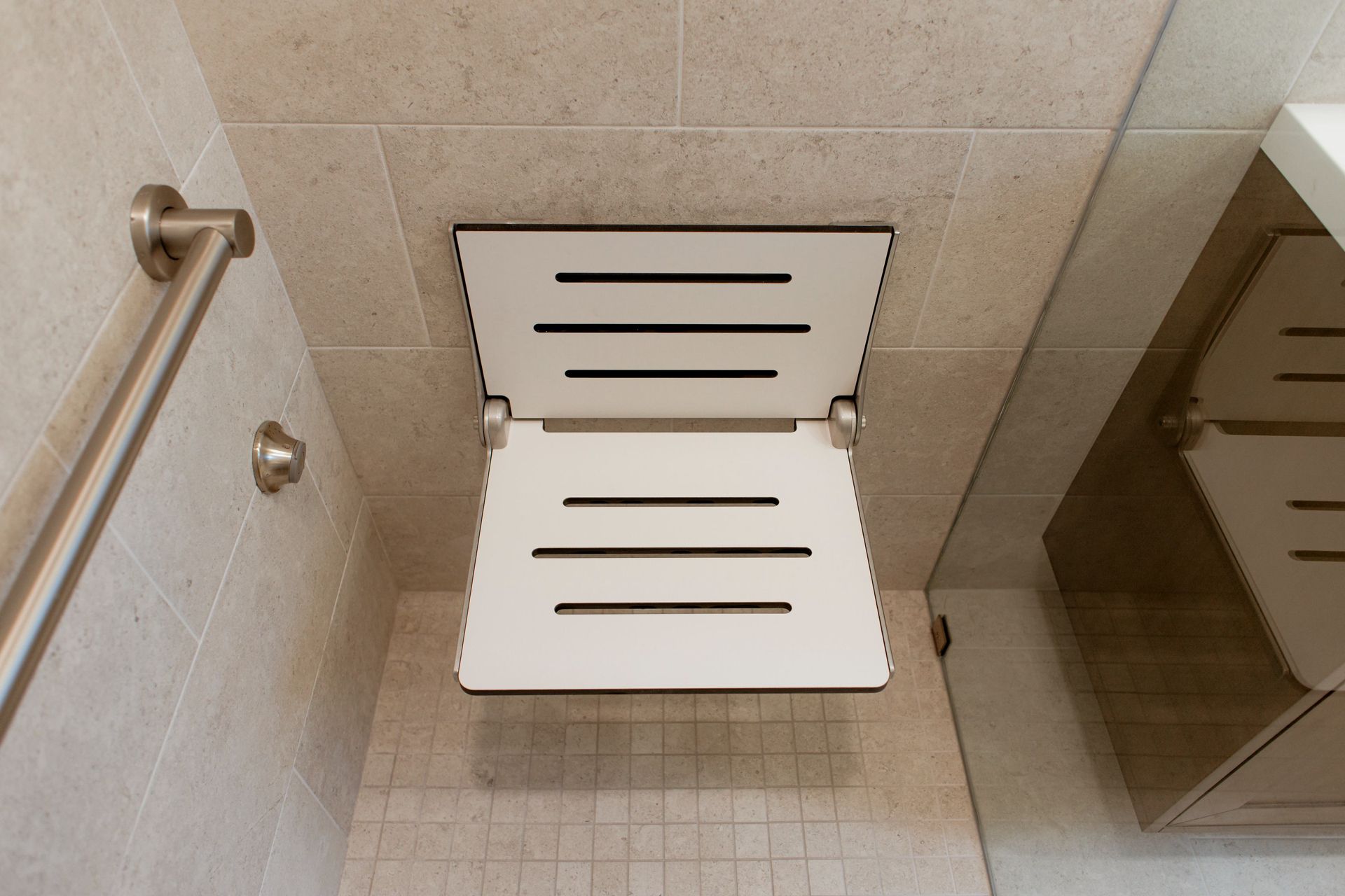 White wall-mounted fold-down seat in a tiled shower stall with grab bar and adjacent partition.