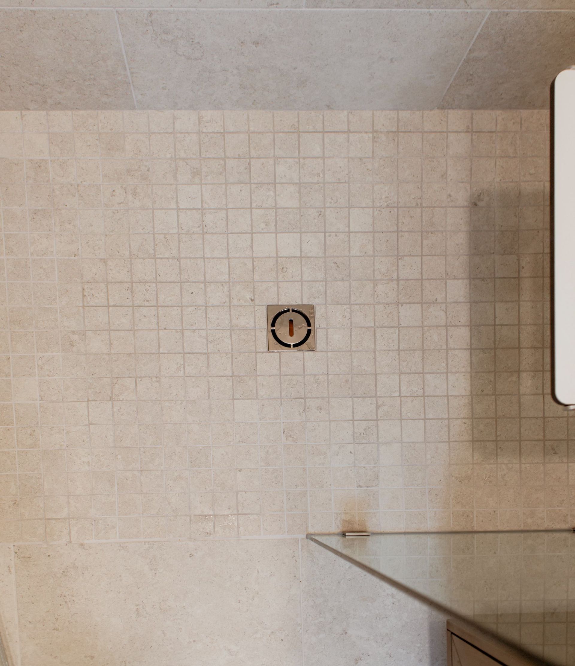 Bathroom tiled wall with a central drain opening above a glass shower shelf.