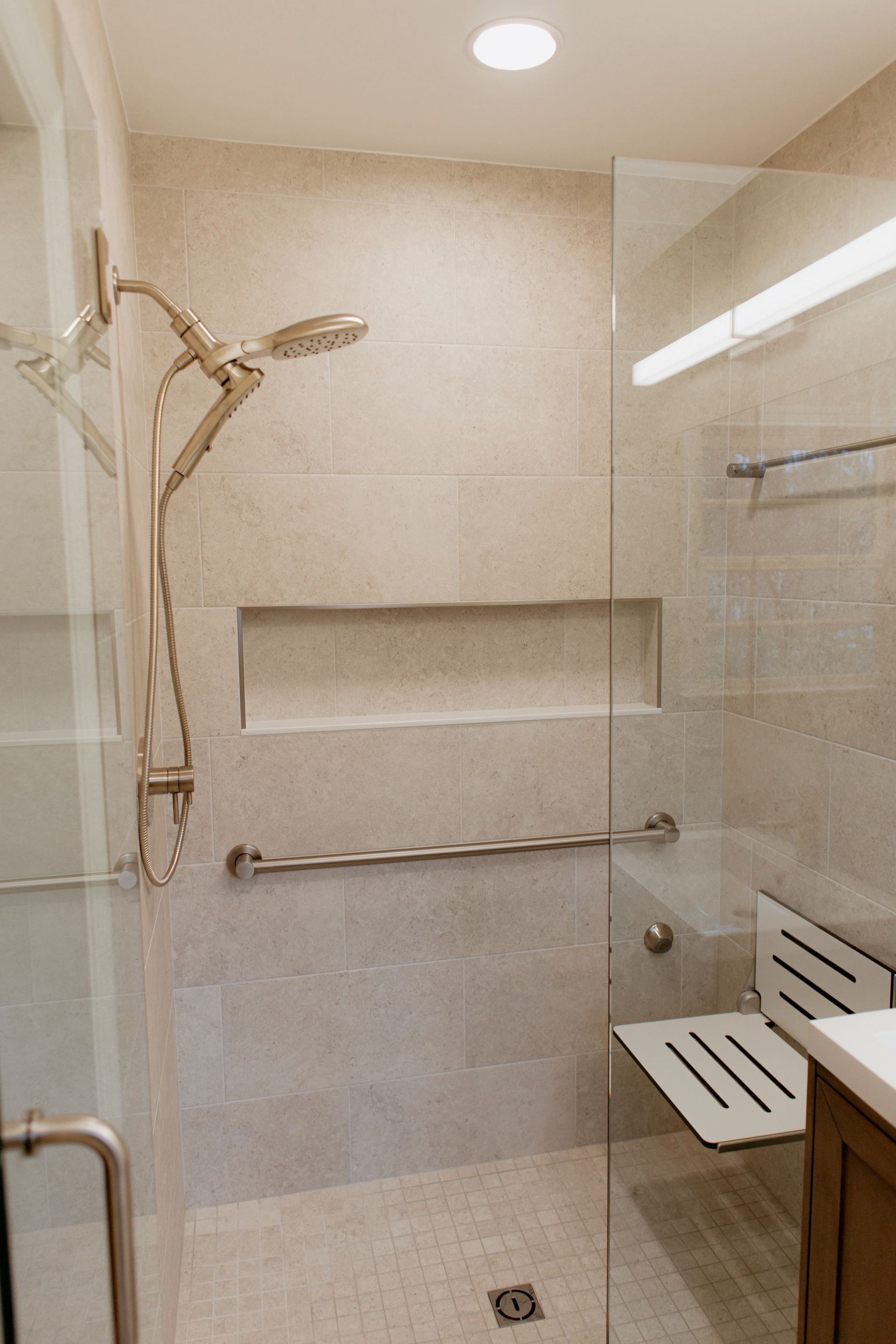 Modern tiled shower with glass door, bench, grab bars, and wall niche under a ceiling light