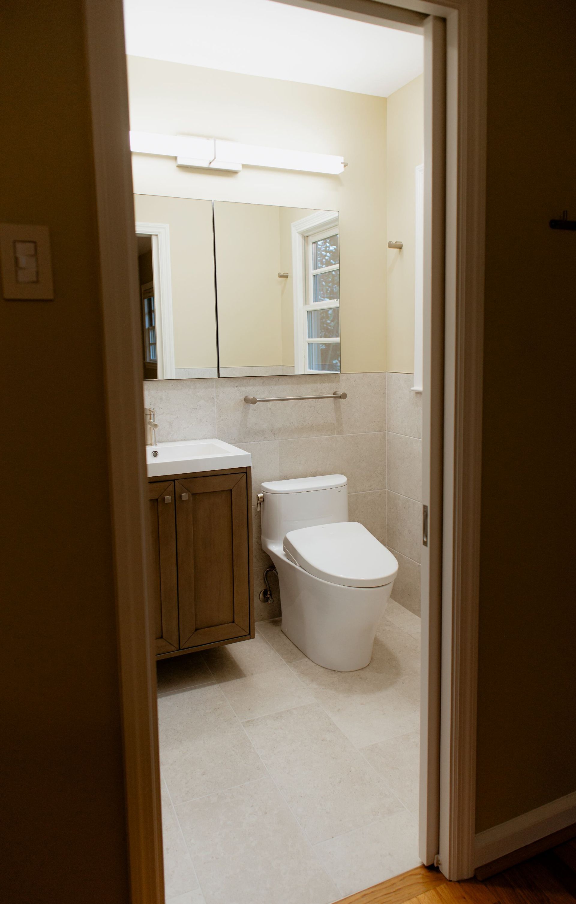 Open doorway to a small bathroom with a toilet, sink, and mirror, lit by bright overhead lights.
