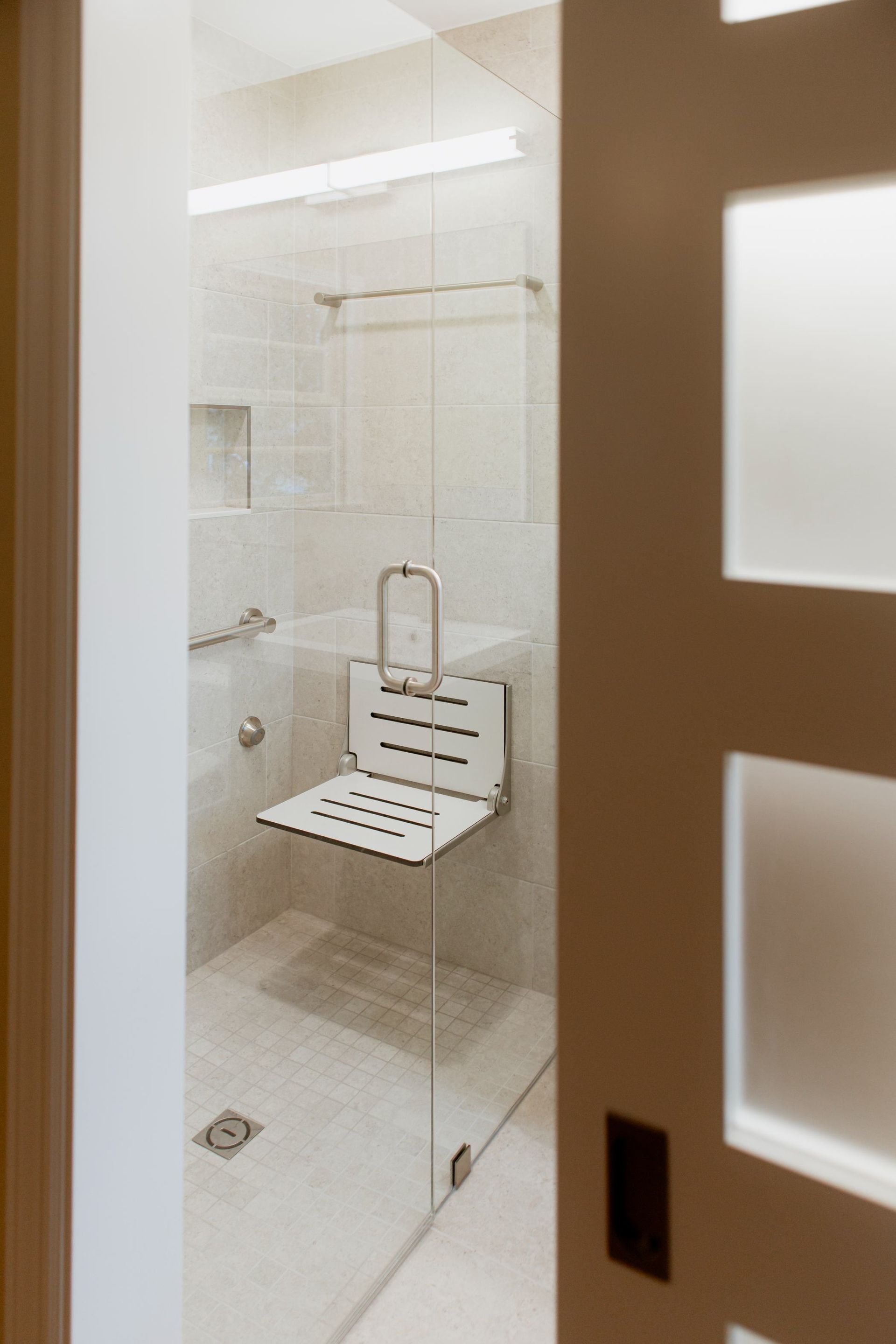 Glass shower stall in a bright tiled bathroom, with a white fold-down seat and grab bar.