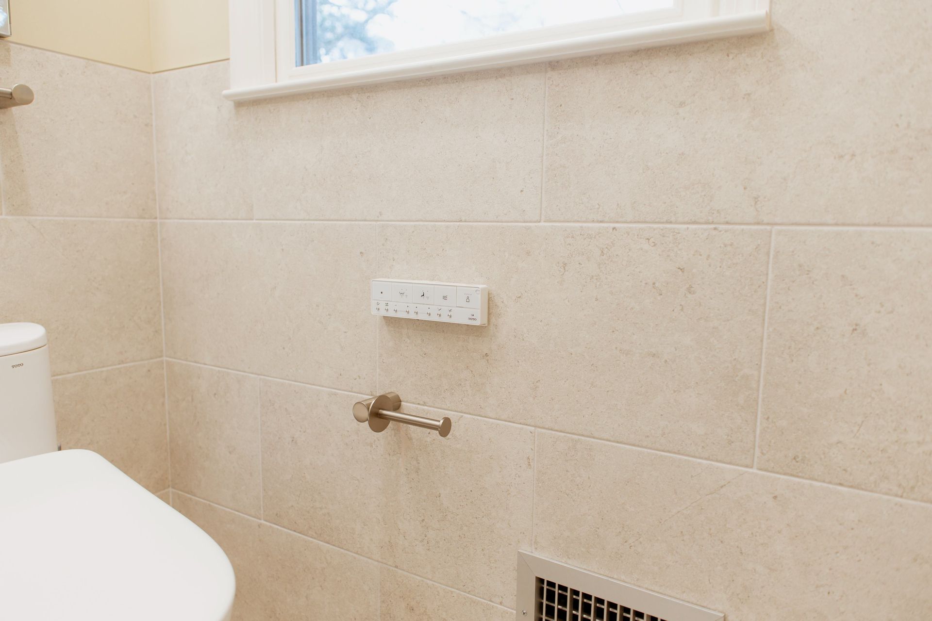 Minimal bathroom wall with toilet, towel hook, vent, and small window above beige tile.
