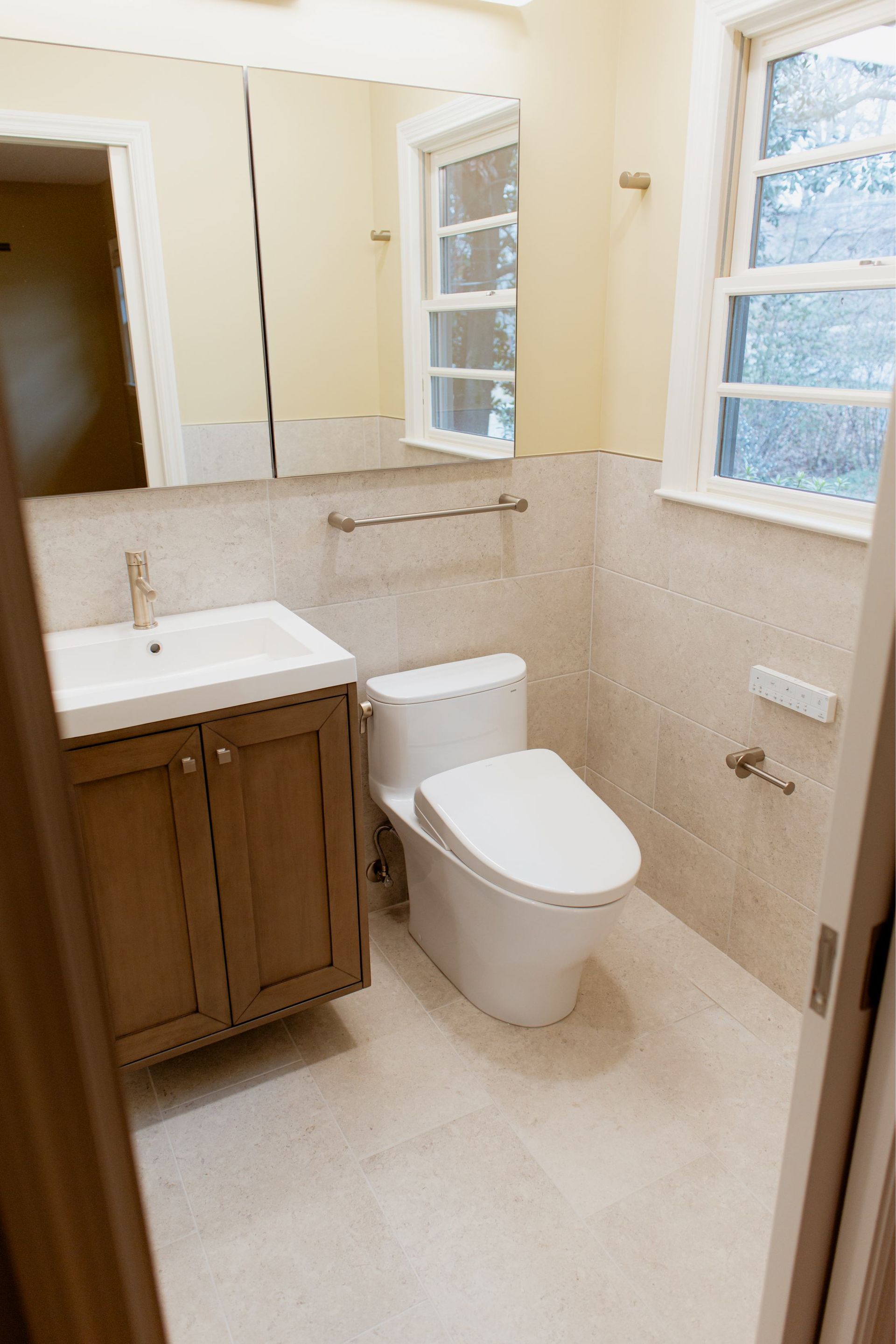 Small beige bathroom with a vanity, toilet, wall mirror, and two windows.