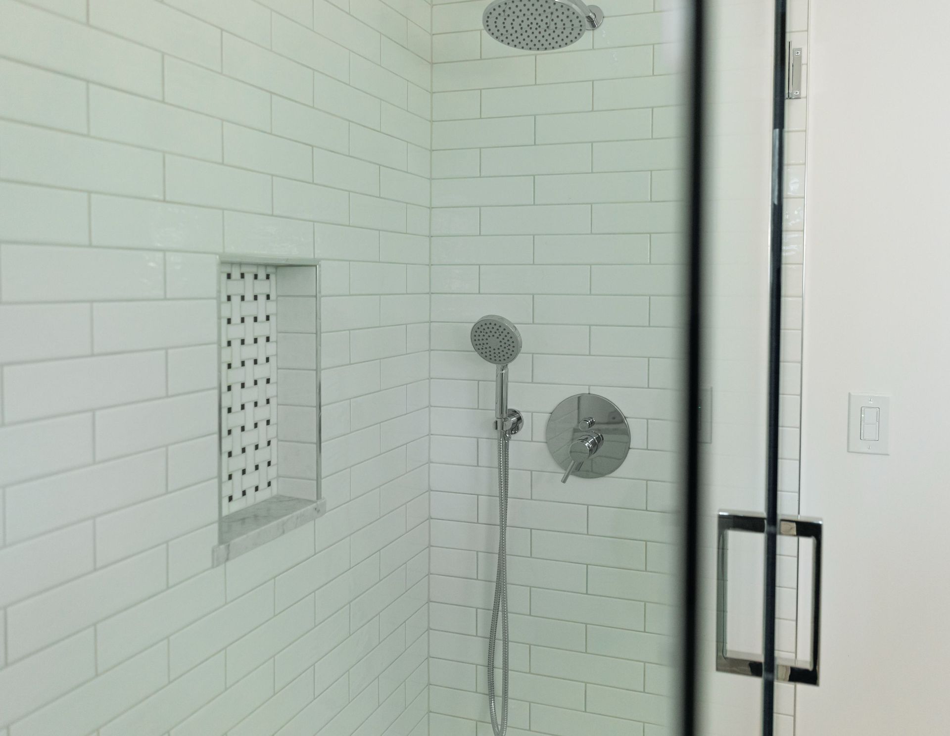 Modern white-tiled shower with rain and handheld showerheads behind a glass door
