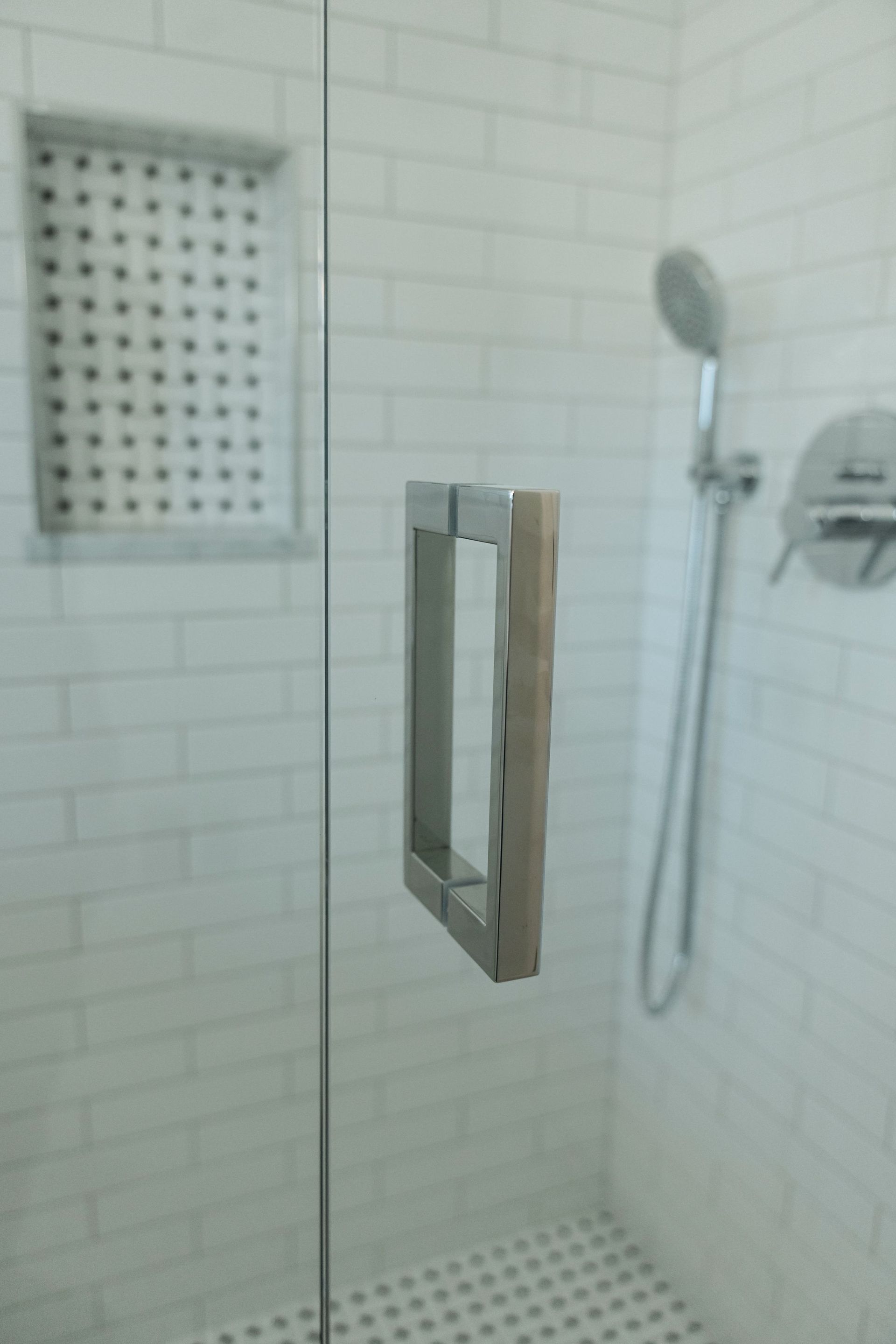 Glass shower enclosure in a white tiled bathroom with a handheld showerhead and wall shelf