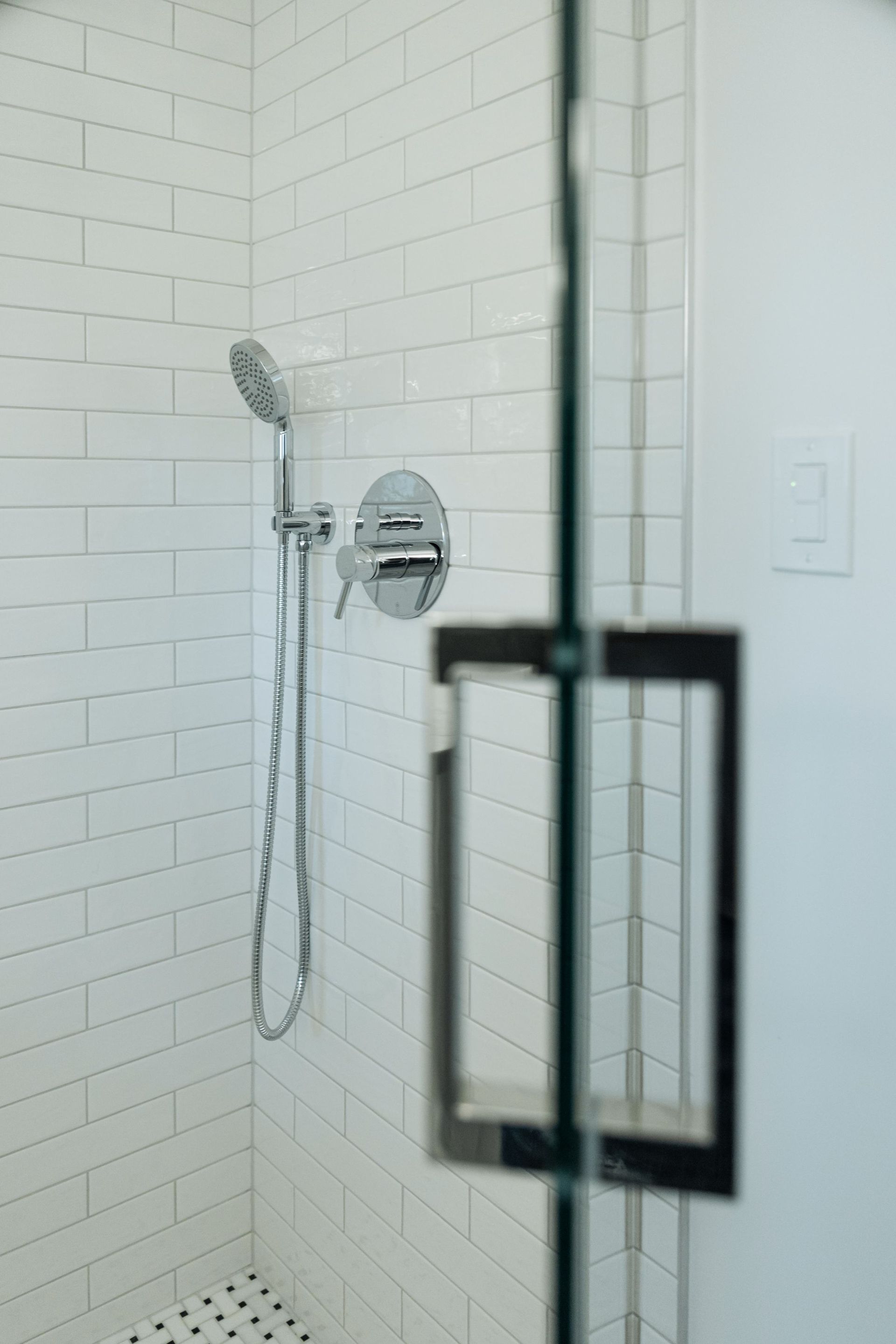 Small white-tiled shower with handheld showerhead, chrome fixtures, and a black-framed glass door panel.