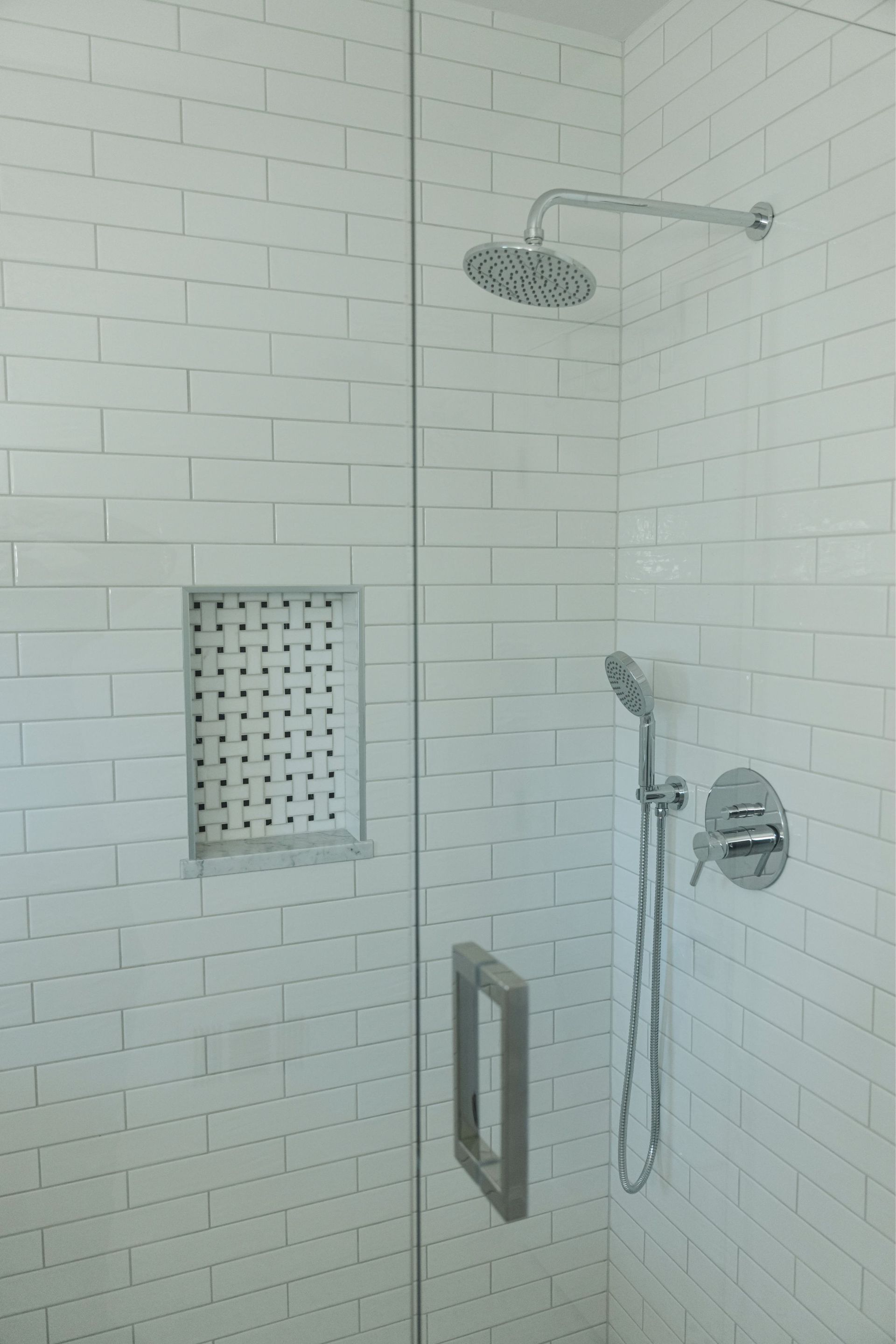 White tiled walk-in shower with showerhead, handheld wand, niche, and grab bar.