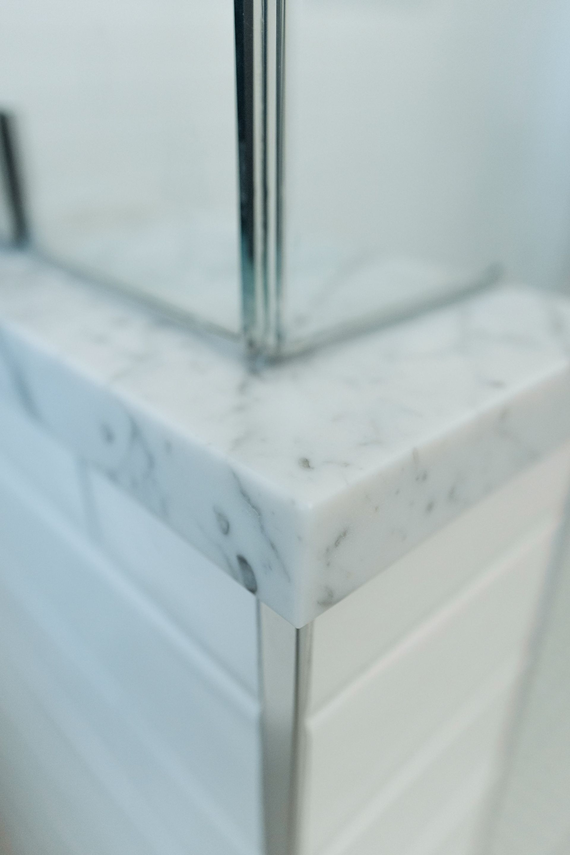 Corner of a white tiled wall with a marble cap and a chrome shower rod