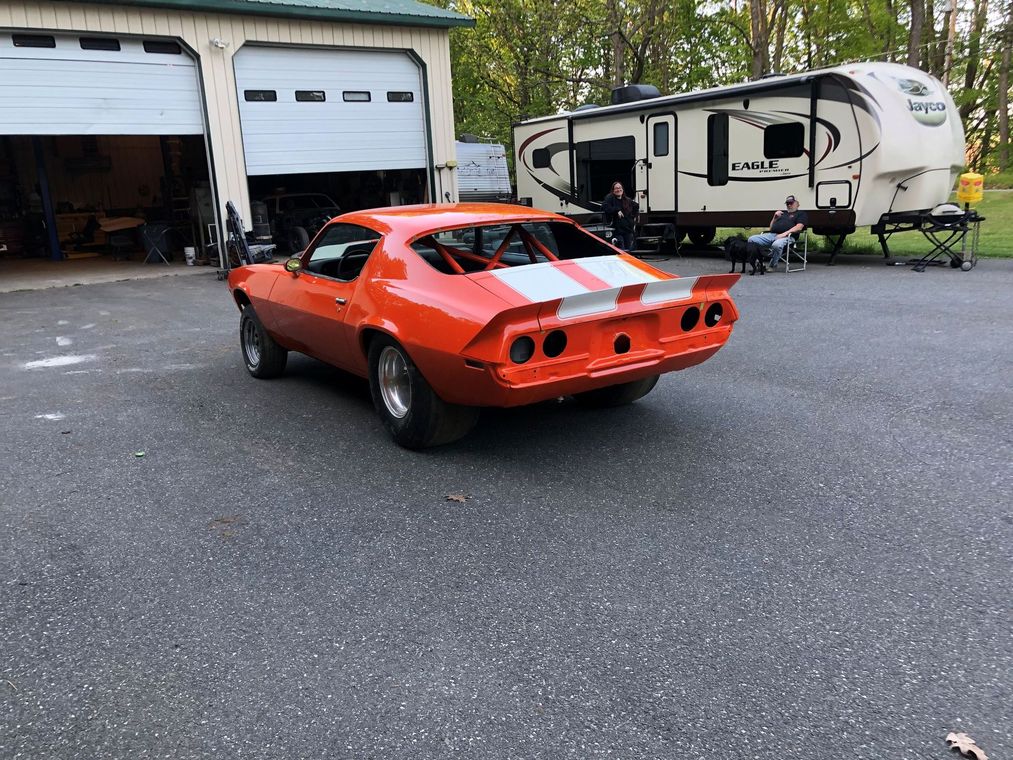 Orange classic muscle car parked by a garage and RV trailer on a gravel lot