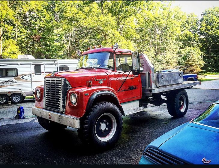 Red vintage flatbed truck parked outdoors beside a blue car and RV in a wooded lot