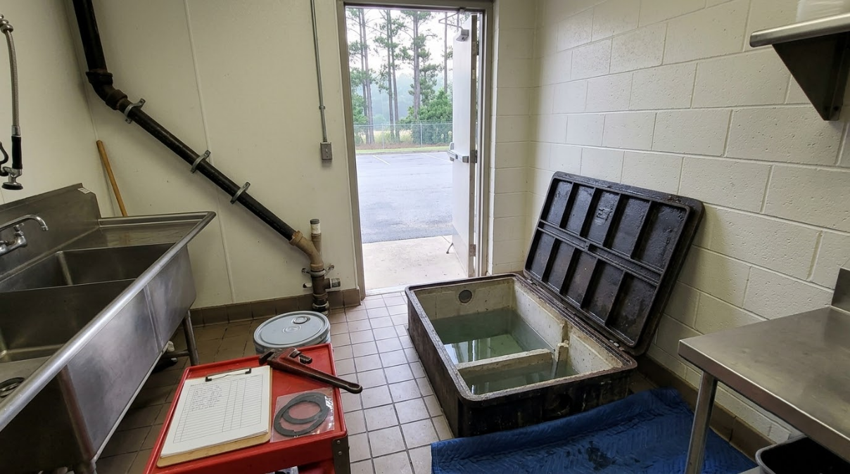 A small tiled utility room with a stainless sink, open containers, and an open door to outside.