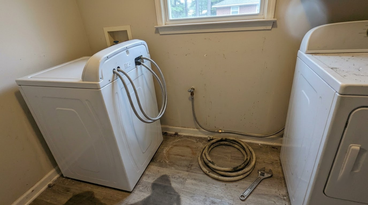 Laundry room with washer and dryer, hoses coiled on the floor by a window.