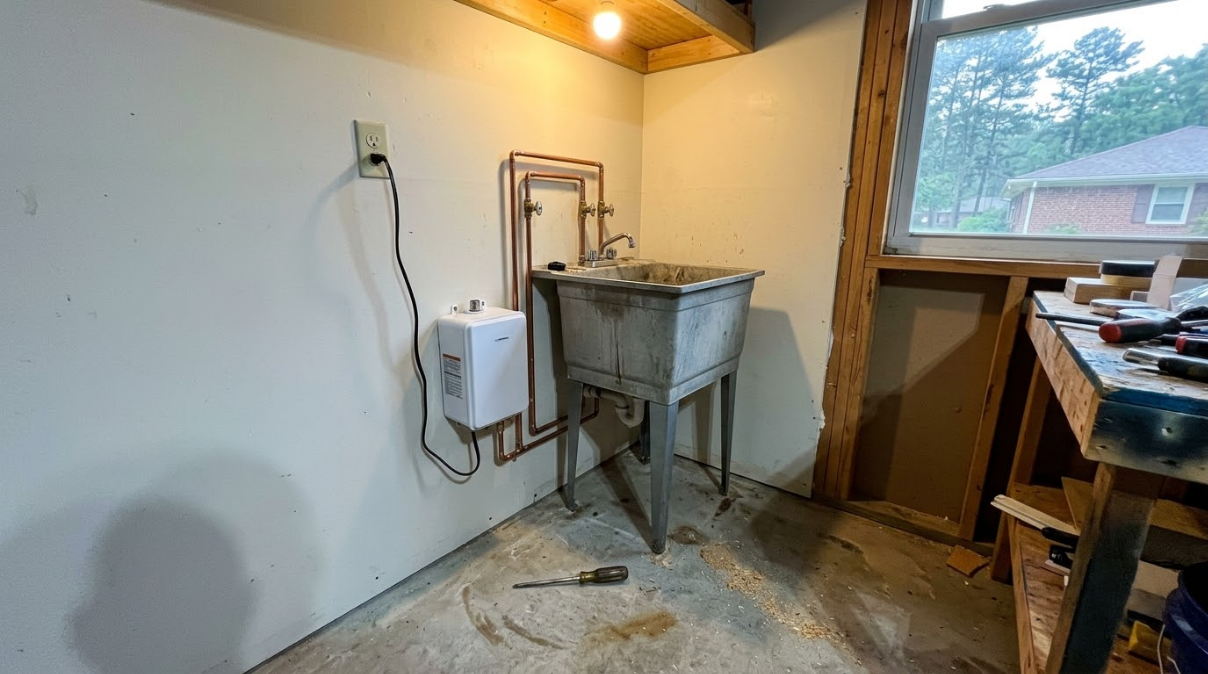 Utility room with a metal sink, wall pipes, and a workbench beside a window.