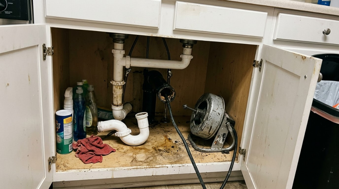 Open under-sink cabinet with exposed plumbing, cleaning spray, rags, and a small fan inside.