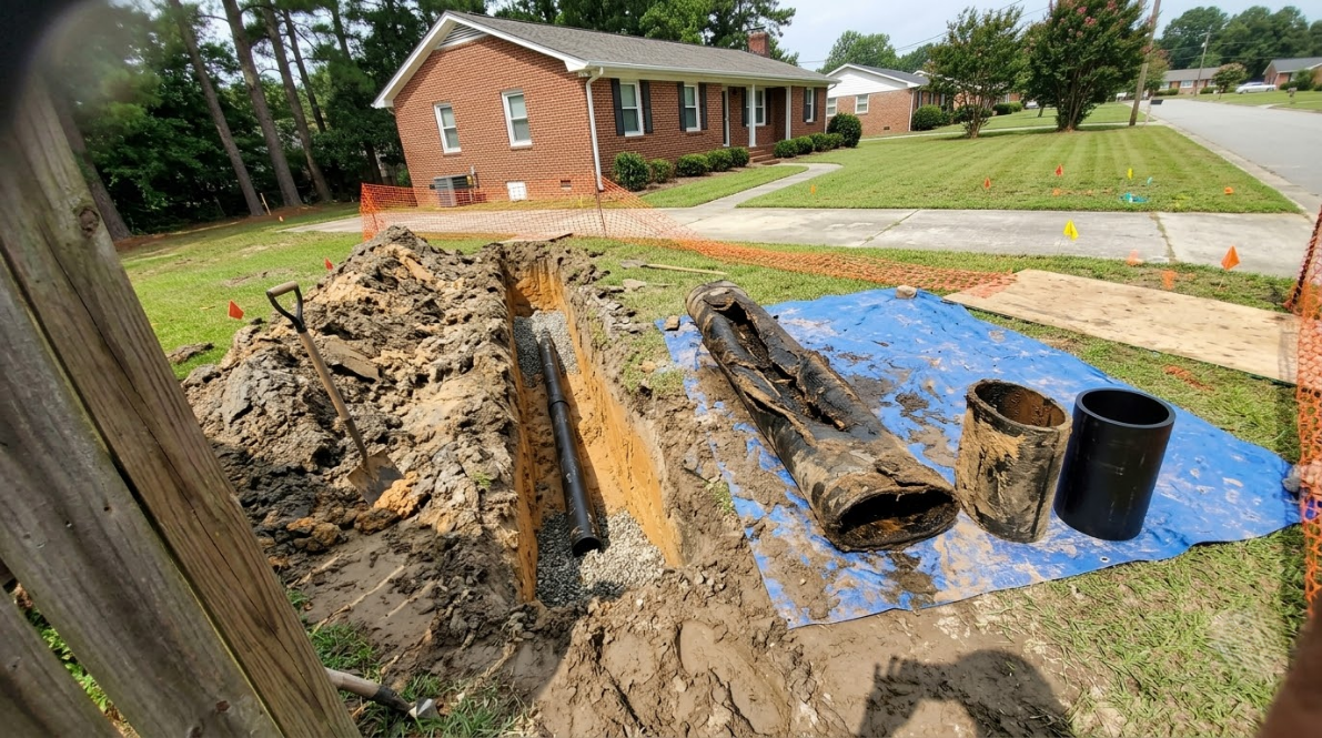 Excavated trench beside brick house with blue tarp, buckets, and muddy soil in a yard