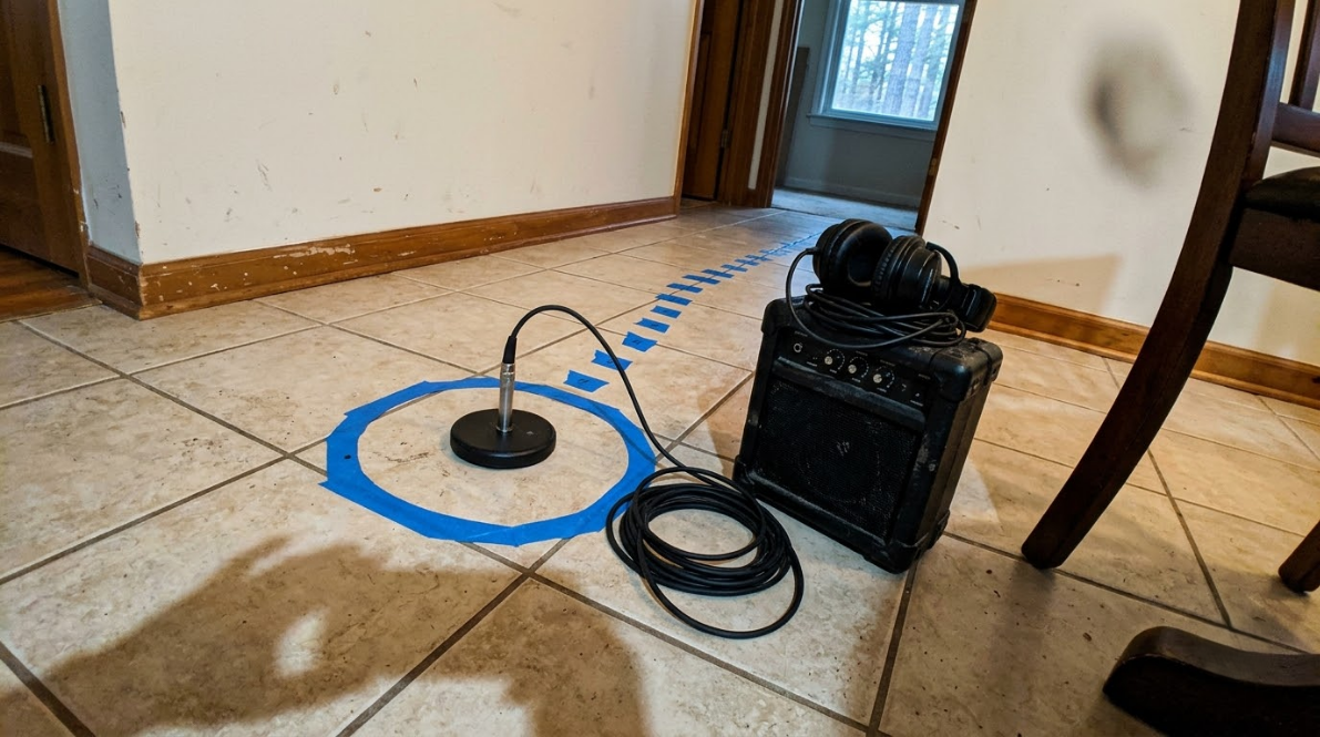 Black speaker with coiled cable on tiled floor beside a blue-taped circle and a small microphone stand