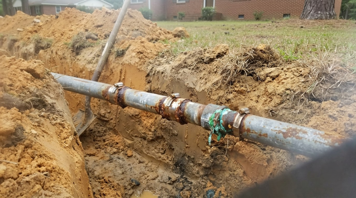 Broken metal pipe in a muddy trench, with water leaking around a rusty joint.