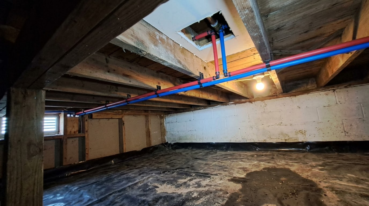 Crawl space under a house with exposed wooden beams, pipes, and a dirt floor.