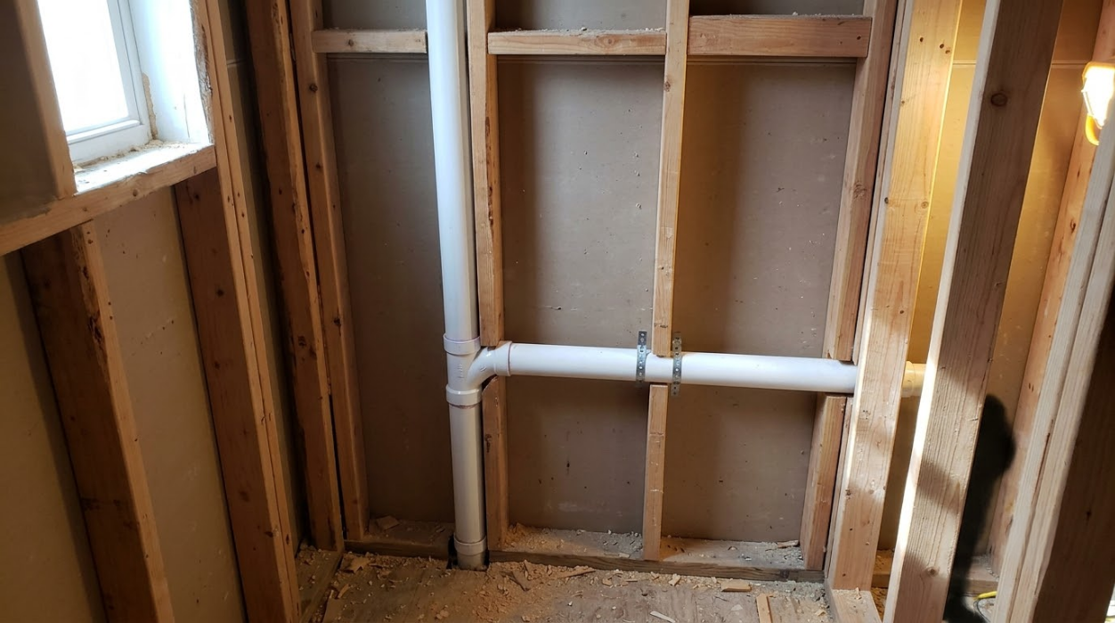 Interior wall framing with exposed studs, white PVC plumbing, and a window at left