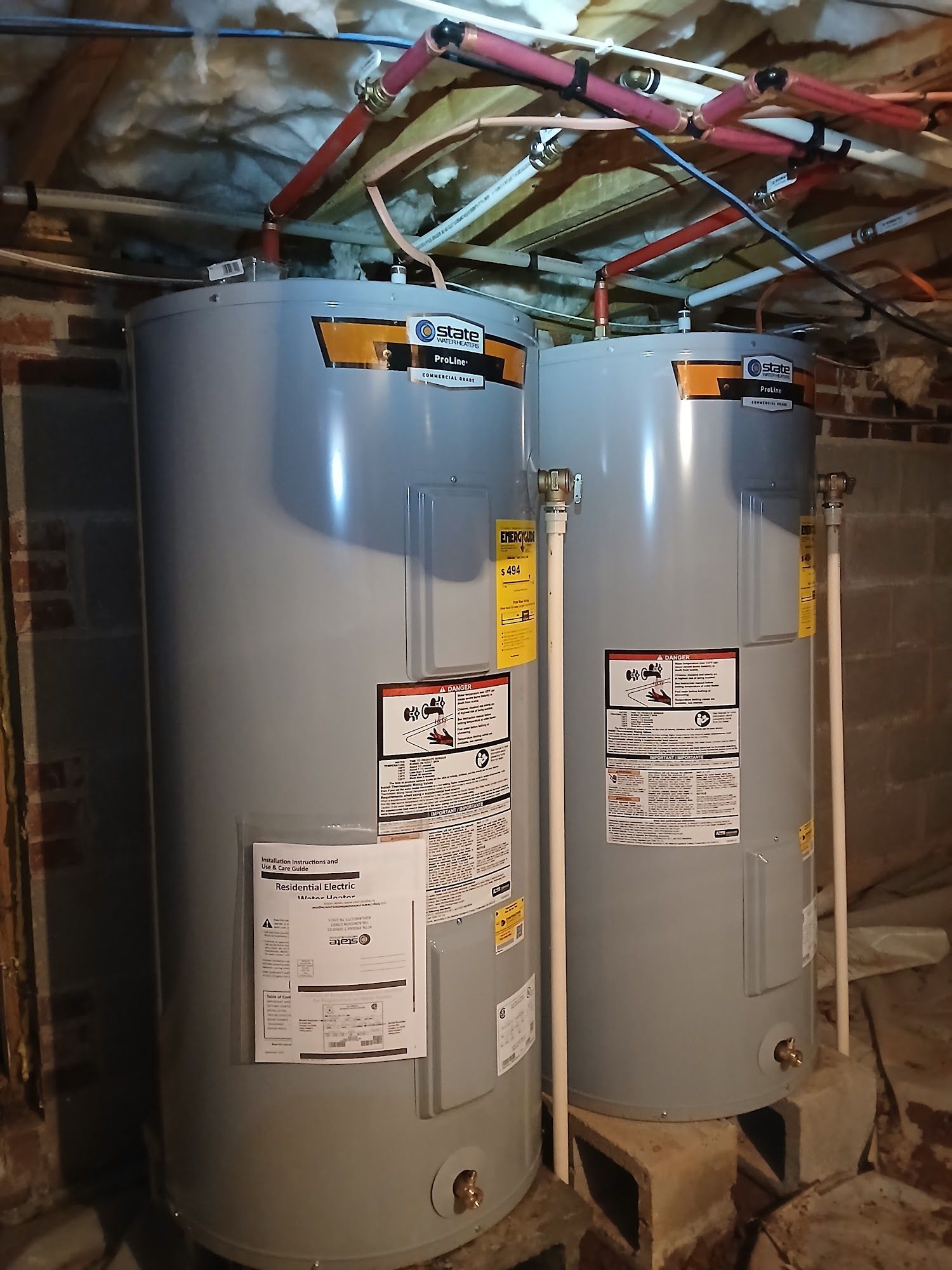 Two gray water heaters in a basement with exposed pipes and ductwork above them