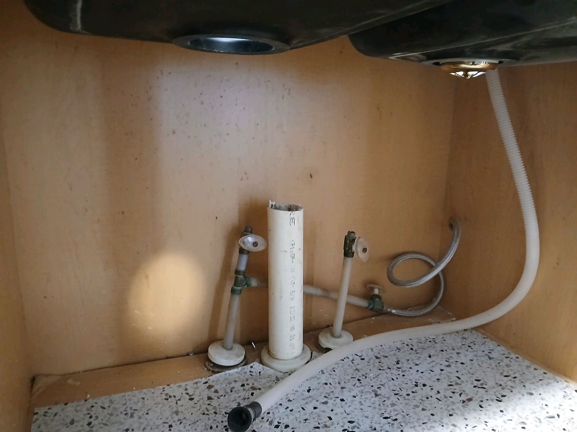 Under-sink cabinet with plumbing pipes, a drain opening, and a flexible water hose on a wood surface.
