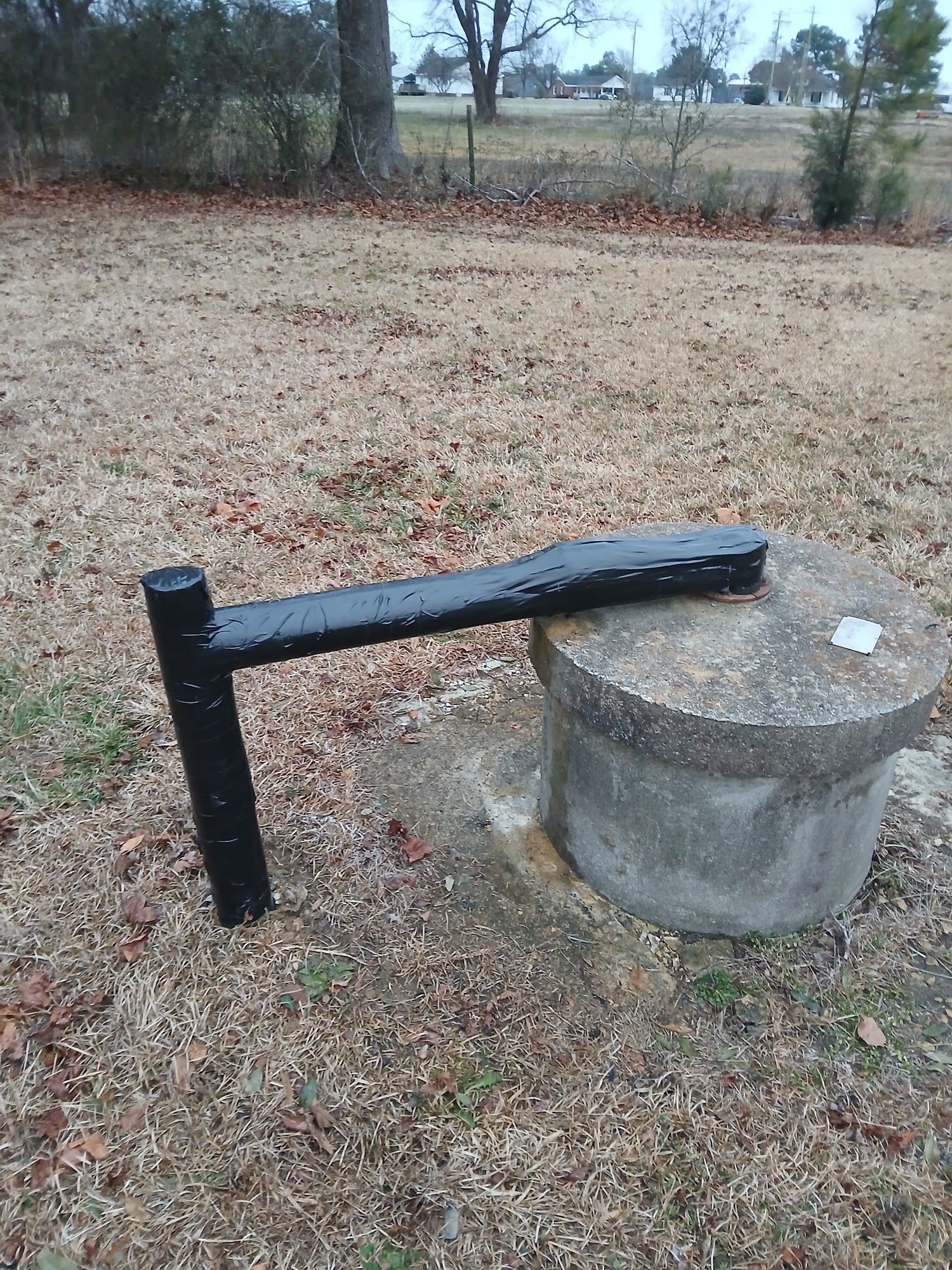 Black metal handle on a concrete well cover in a grassy field with trees in the background