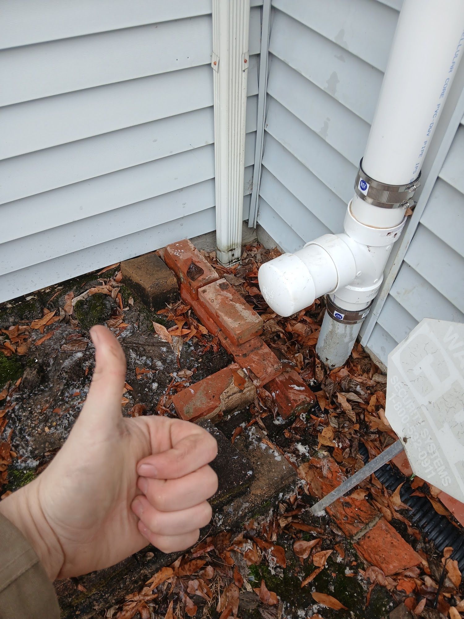 Thumbs-up gesture beside white PVC pipes and a brick-lined corner with fallen leaves outdoors.
