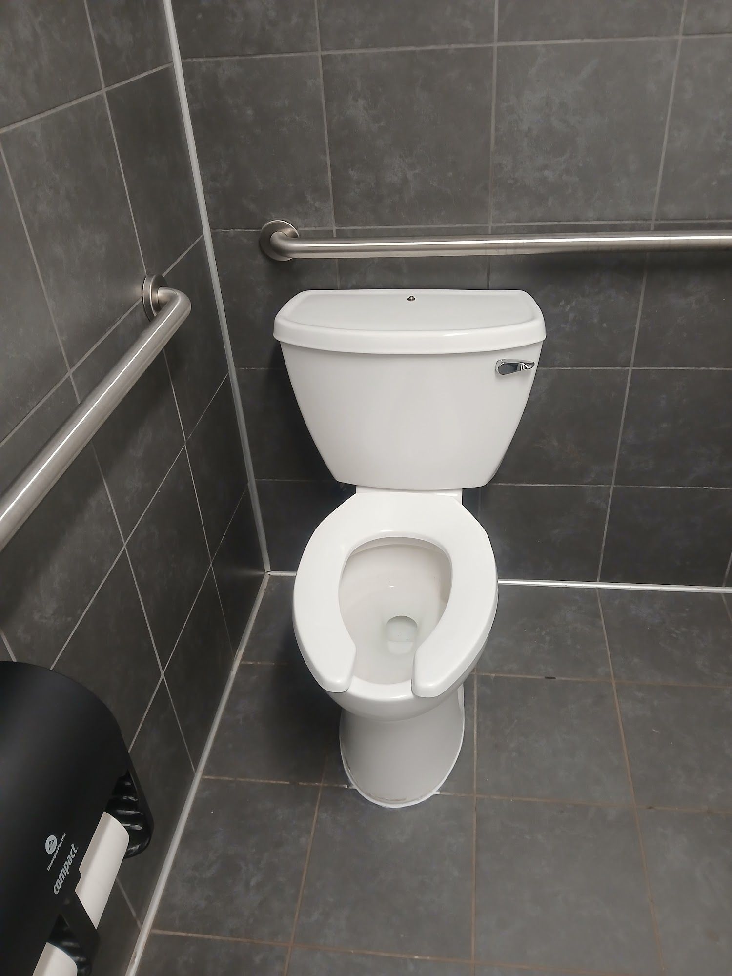 White toilet in a gray-tiled restroom with grab bars on the walls.