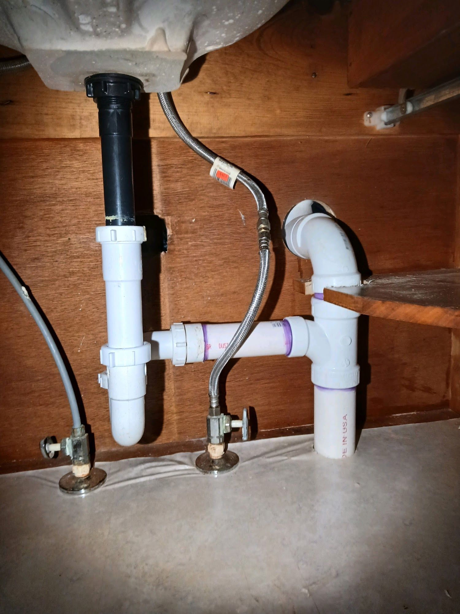 Under-sink plumbing with white PVC pipes and a chrome faucet hose against a wood cabinet wall
