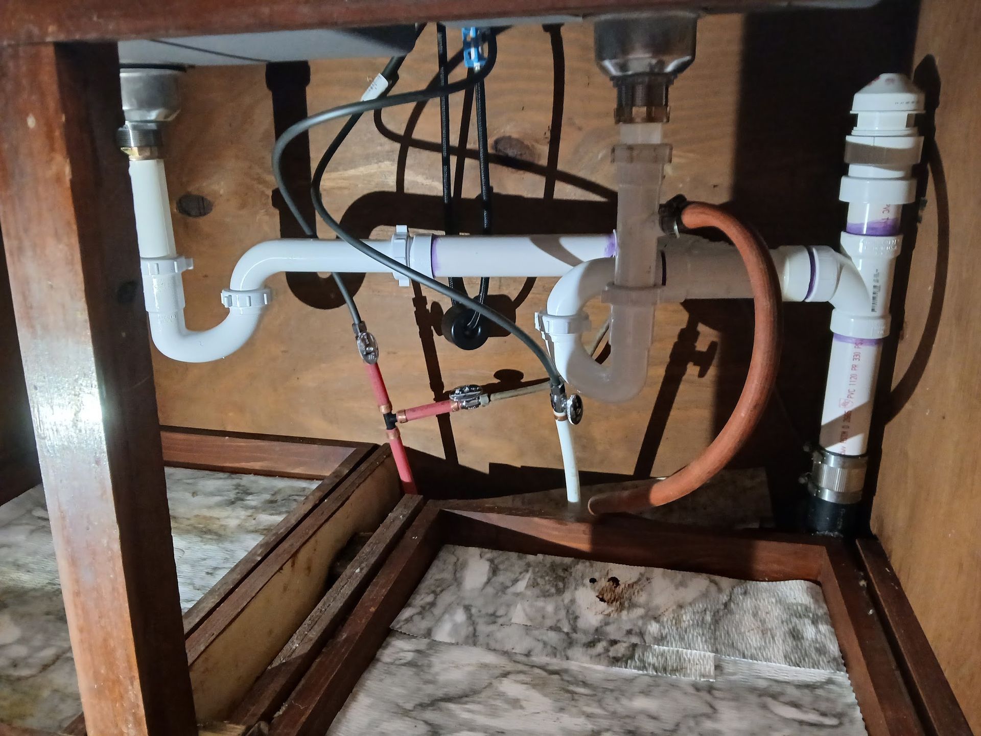 Under-sink plumbing with exposed pipes, valves, and a marble-pattern cabinet base opening