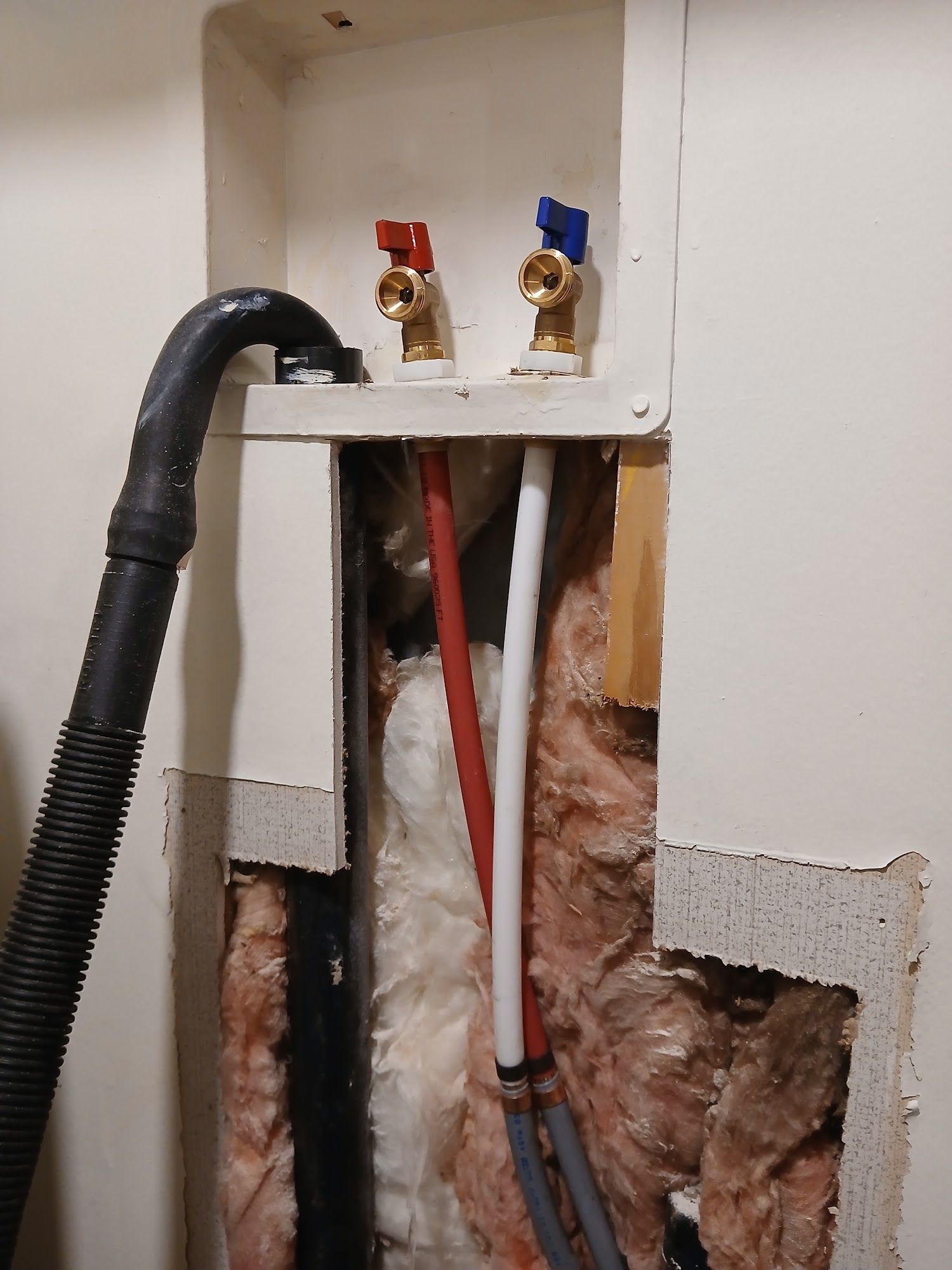 Exposed plumbing and wiring inside a wall opening with red and white pipes and brass valves