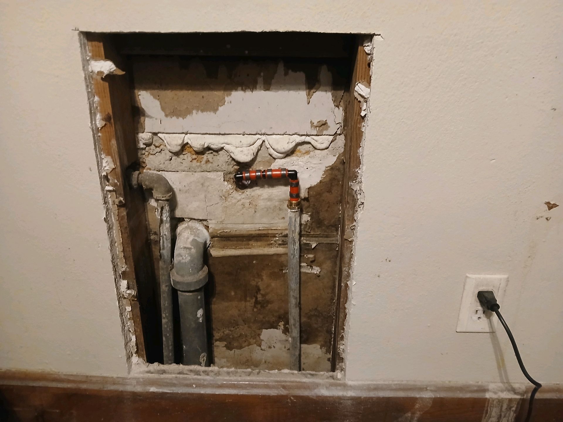 Open wall with exposed pipes and plumbing in a beige room, near an electrical outlet and cord.
