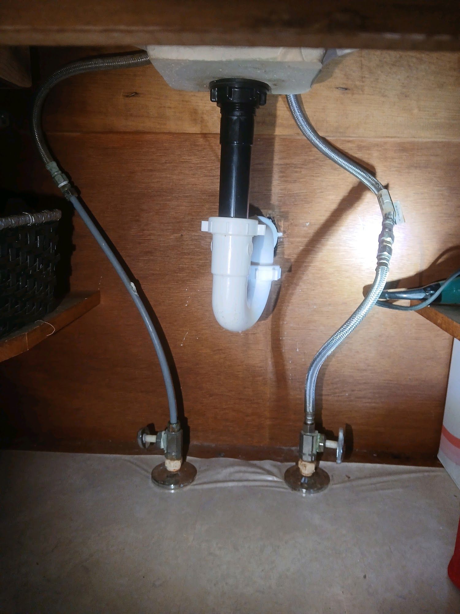 Under-sink plumbing with a white P-trap and two water supply lines under a wooden cabinet