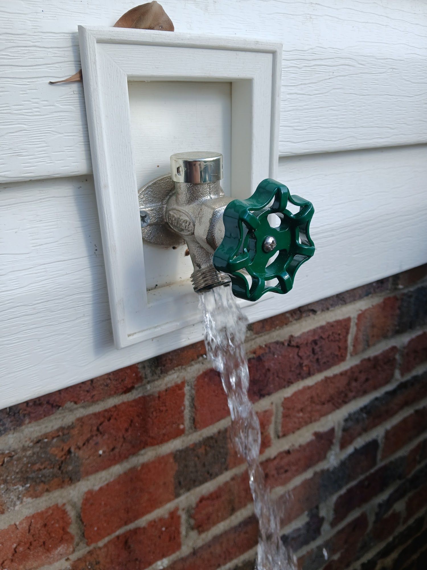 Outdoor water spigot with a green valve handle mounted on a brick wall, water running out