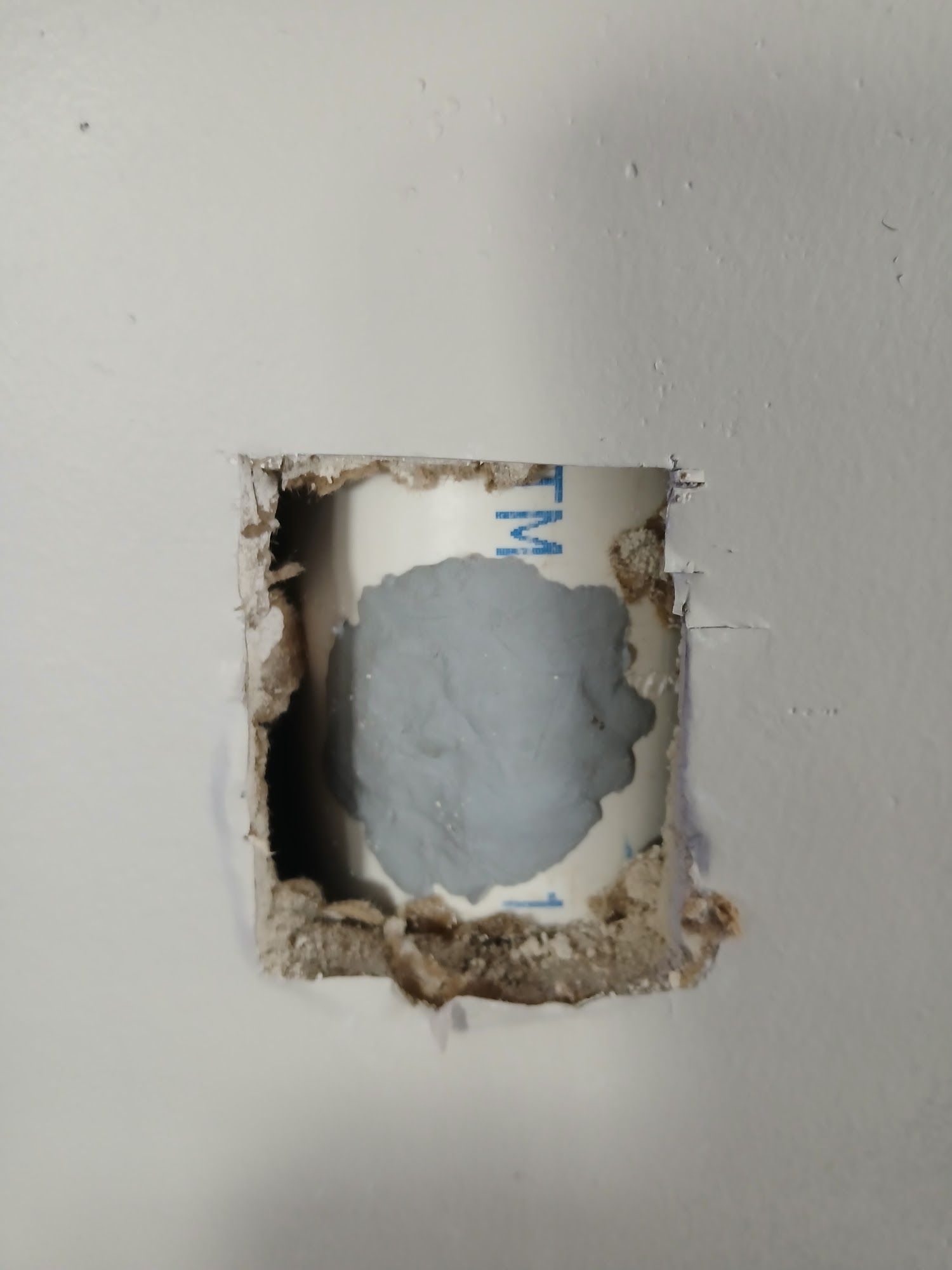Square hole cut in a white wall, exposing a gray pipe or duct behind it.