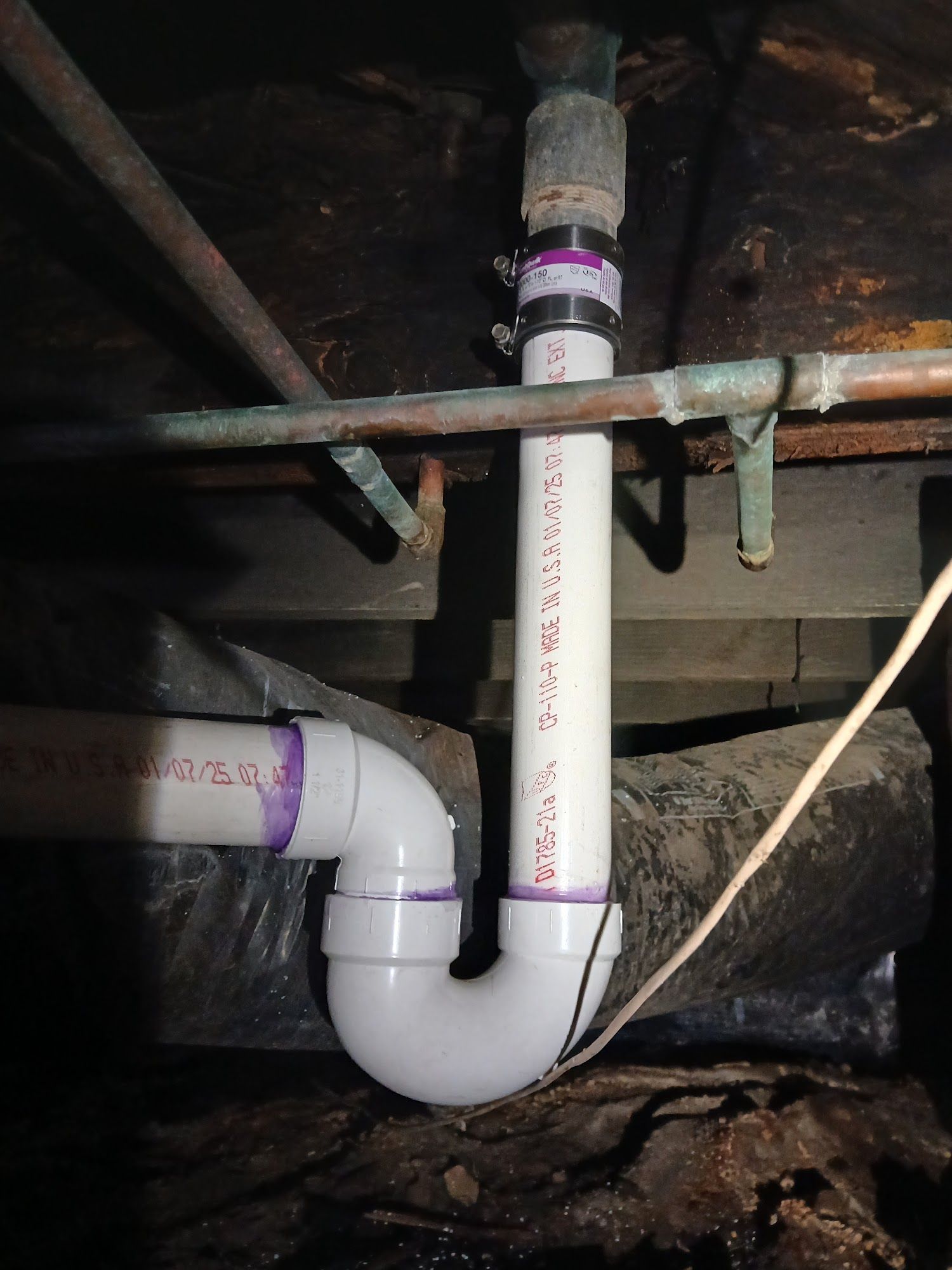 Under-sink plumbing with white PVC pipes and a purple band around a vertical pipe beneath wooden floor joists