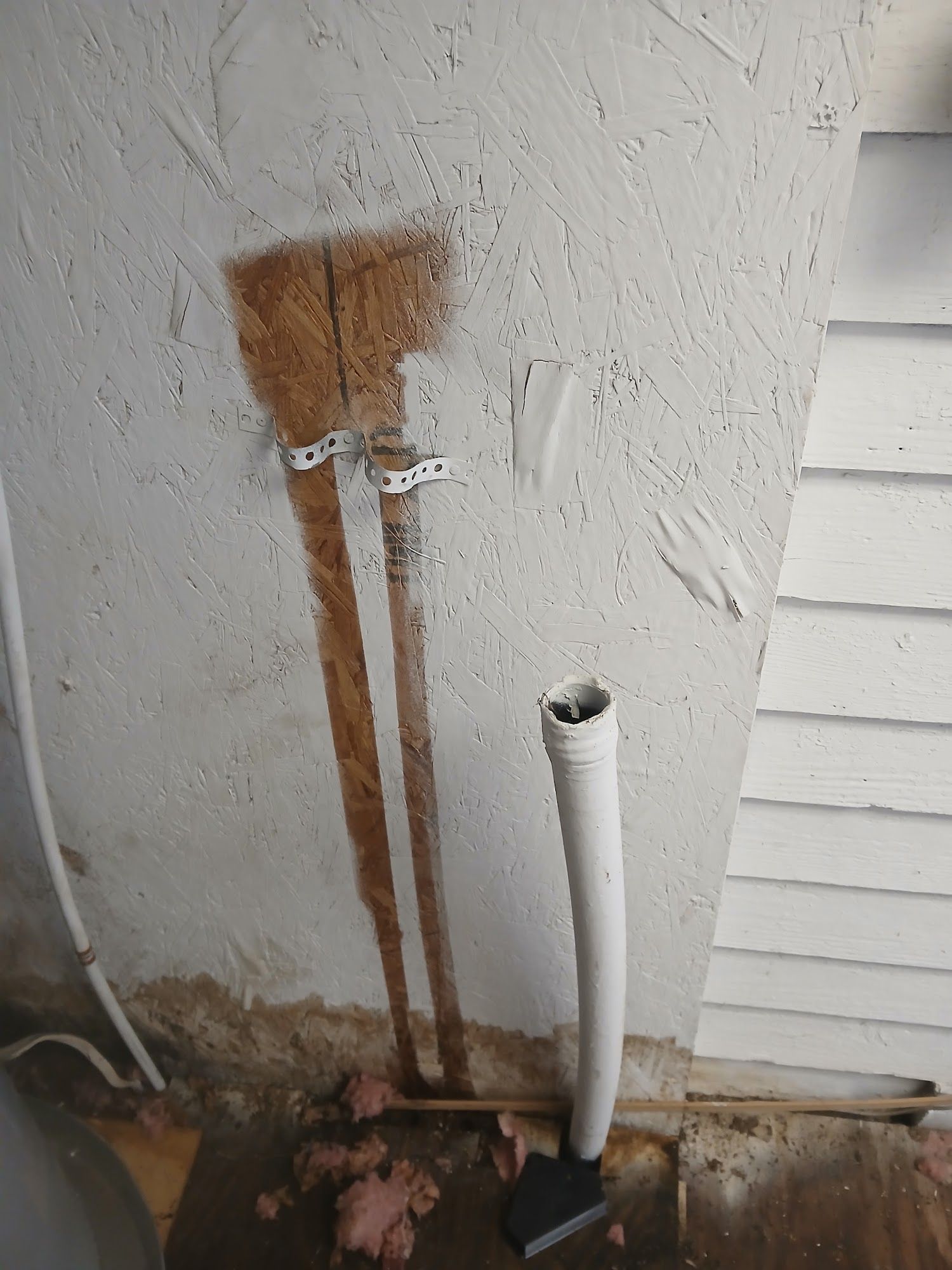 Two rusty metal pipes and a white PVC pipe against a stucco wall with dirt at the base