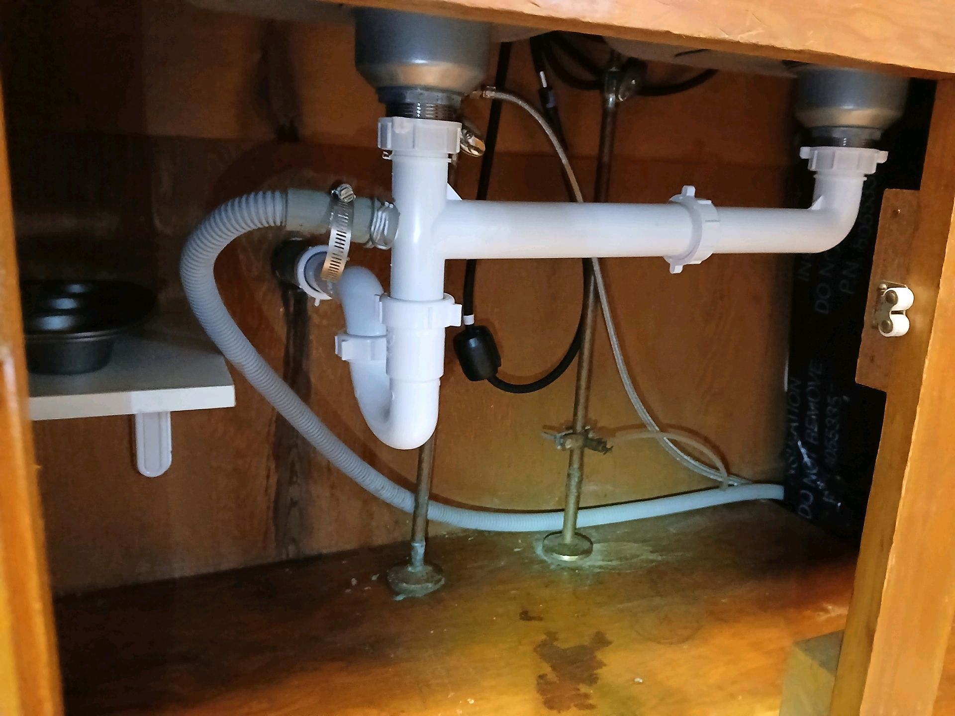 Under-sink plumbing with white pipes, hoses, and a small water stain on the cabinet floor