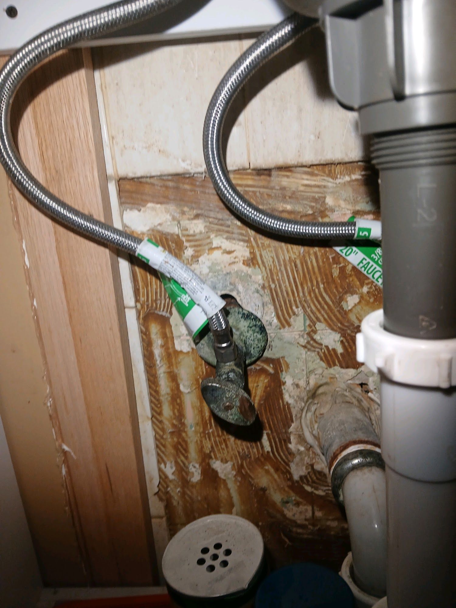 Under-sink plumbing with two braided hoses and a gray drain pipe against a damaged wooden cabinet wall