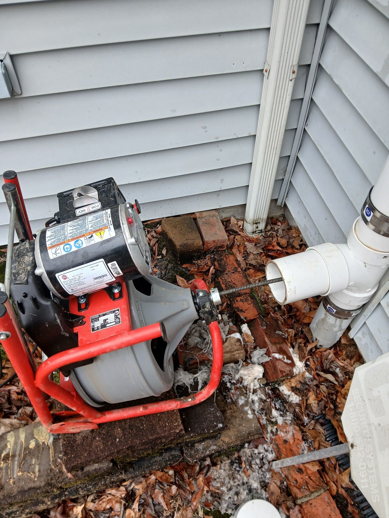 Red drain snake machine beside white PVC pipe and house siding, on muddy leaves outdoors