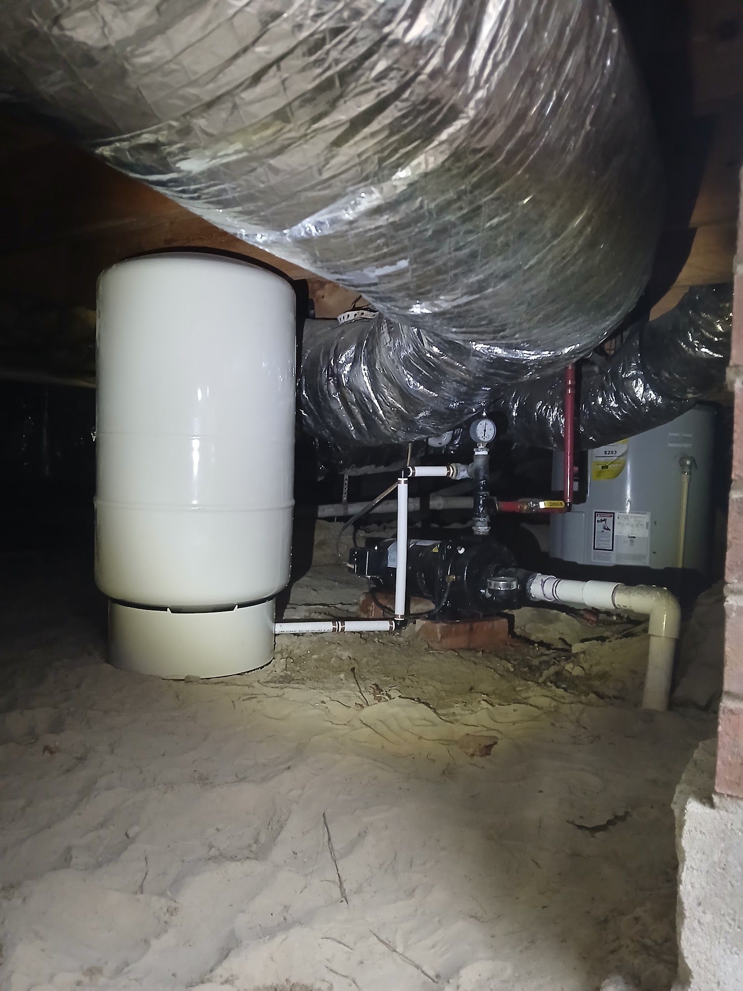 Crawlspace with large silver ductwork, white HVAC tank, pipes, and sandy floor under a house