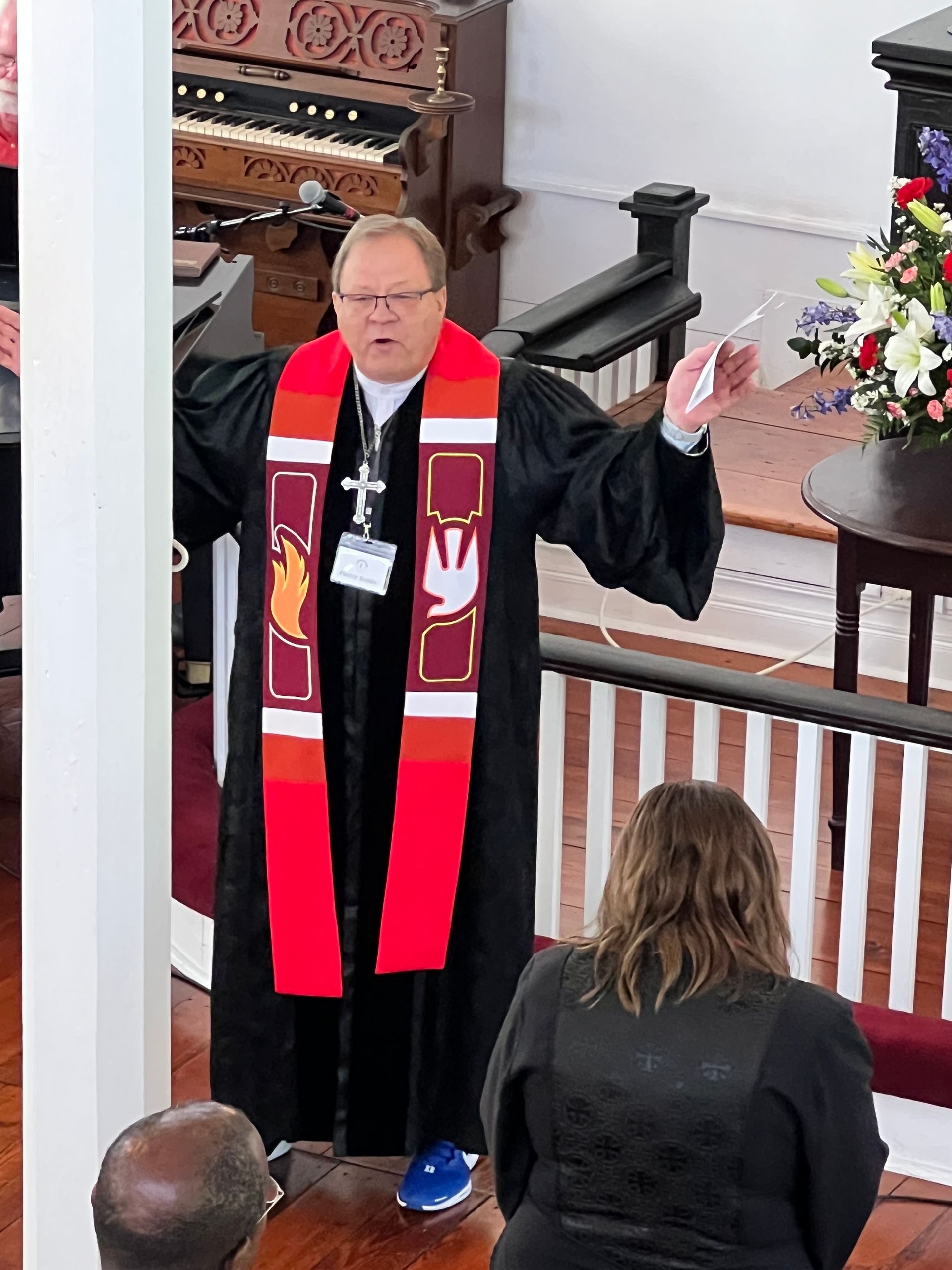 Pastor in black robe, red and maroon stole, preaching in a church.  He has hands raised, a microphone, and a badge.