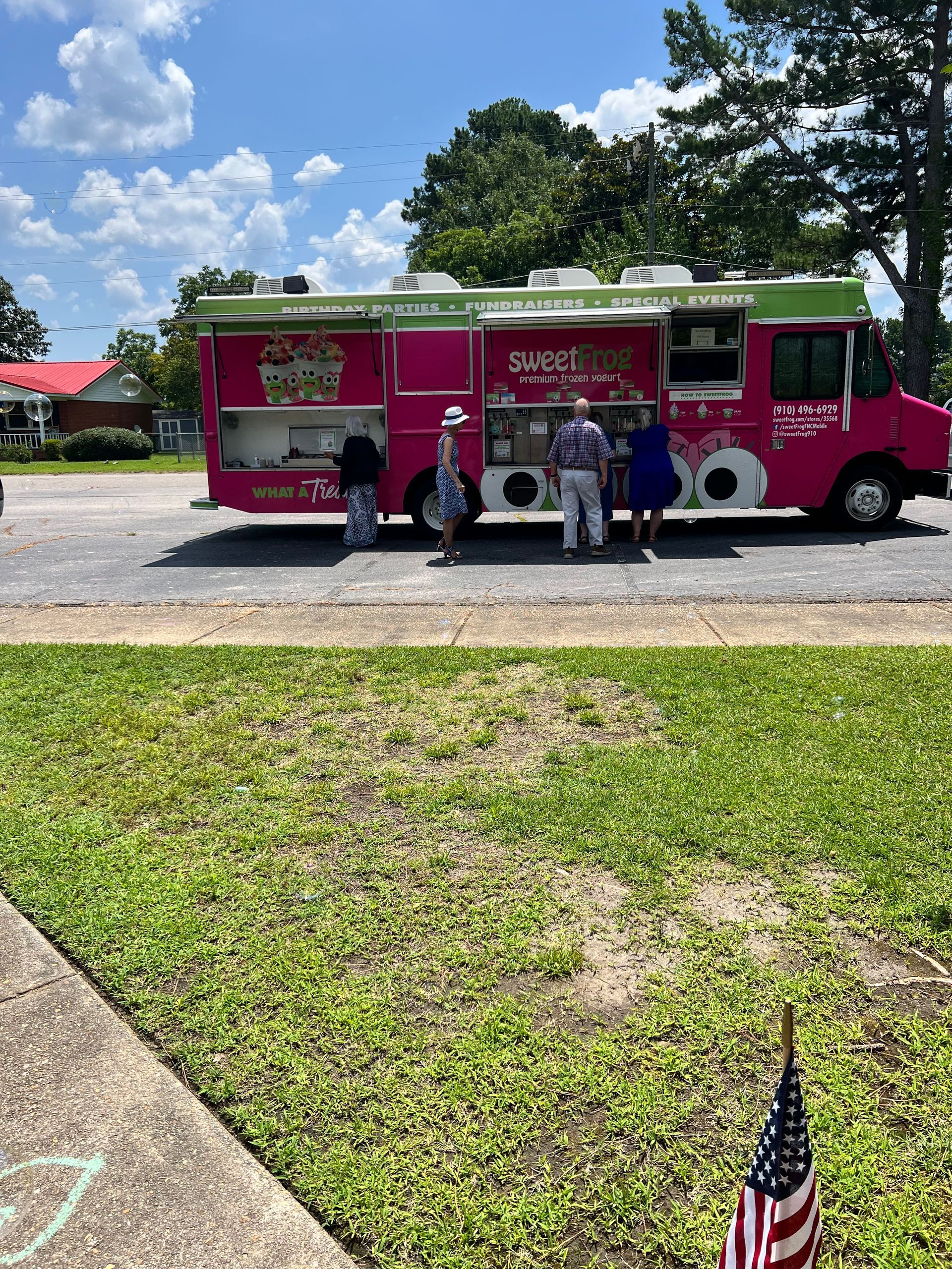Pink food truck with customers. Green grass and American flag in foreground.